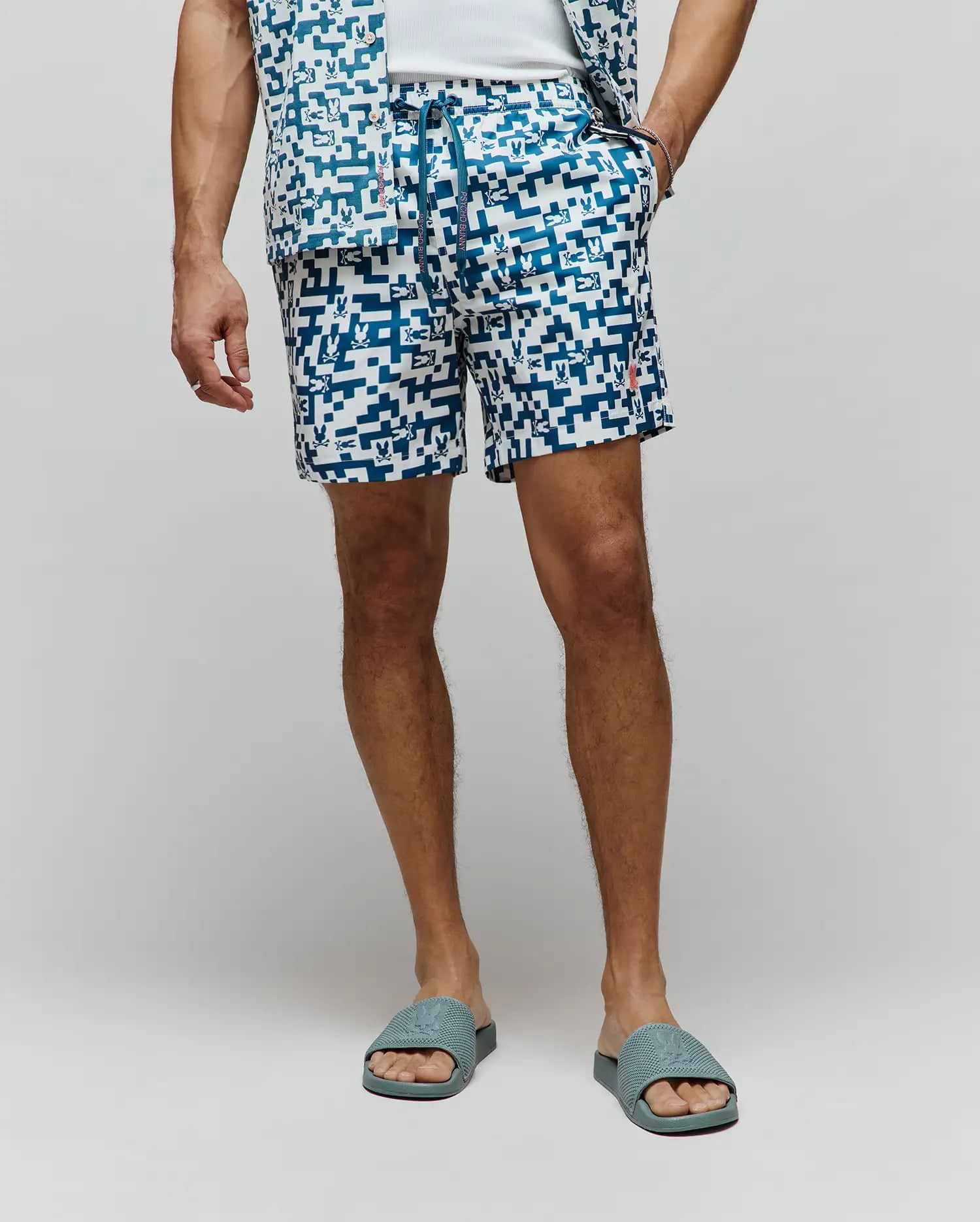 Mens Josh Swim Trunk in white with a teal pixelated geometric pattern featuring small Psycho Bunny skull logos, a branded drawstring, and a small embroidered red bunny logo on the left leg.