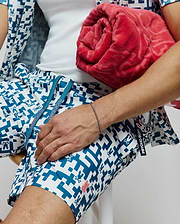 Mens Josh Swim Trunk, classic fit, white with a bold teal geometric pattern incorporating small bunny skull logos. Features teal drawstrings with pink Psycho Bunny text, a pink embroidered bunny on the leg, and a branded side tag.