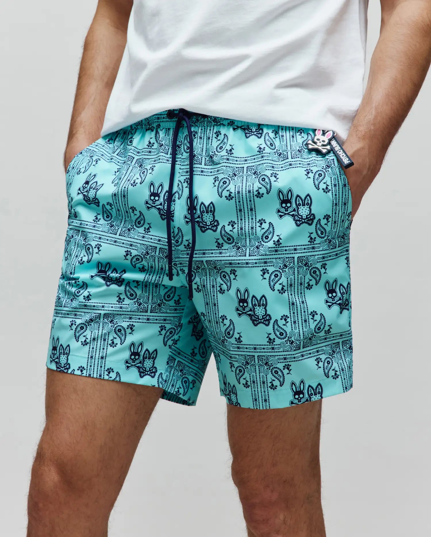 Mens Luke Swim Trunk: Aqua. Navy bandana paisley print with Psycho Bunny skull logos, drawstring, side pockets, and a Psycho Bunny charm with a pink nose on the right pocket.
