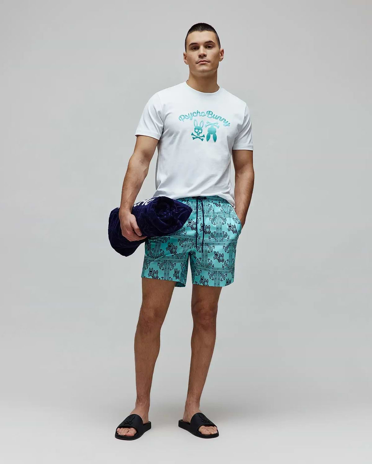 White classic fit jersey t-shirt with a large gradient blue Psycho Bunny text and bunny skull chest graphic. Mens Luke Swim Trunk in teal features an all-over dark blue bandana print with integrated bunny skull logos.