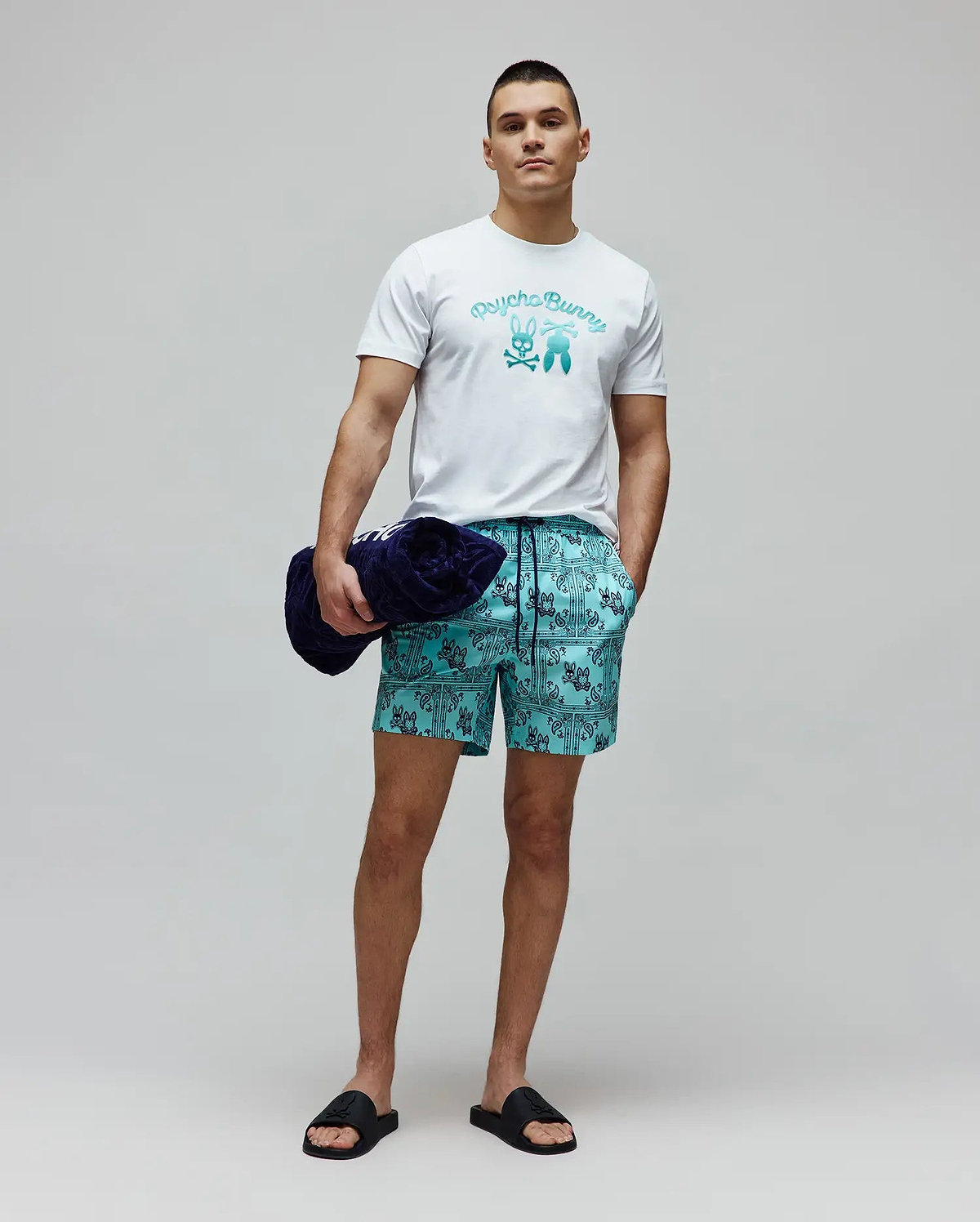 White classic fit jersey t-shirt with a large gradient blue Psycho Bunny text and bunny skull chest graphic. Mens Luke Swim Trunk in teal features an all-over dark blue bandana print with integrated bunny skull logos.