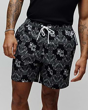 Mens Luke Swim Trunk in black with a white paisley bandana print featuring repeated Psycho Bunny skull logos, a white drawstring, and a Psycho Bunny branded tag. Relaxed fit, smooth fabric.