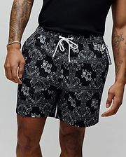 Mens Luke Swim Trunk in black with a white paisley bandana print featuring repeated Psycho Bunny skull logos, a white drawstring, and a Psycho Bunny branded tag. Relaxed fit, smooth fabric.