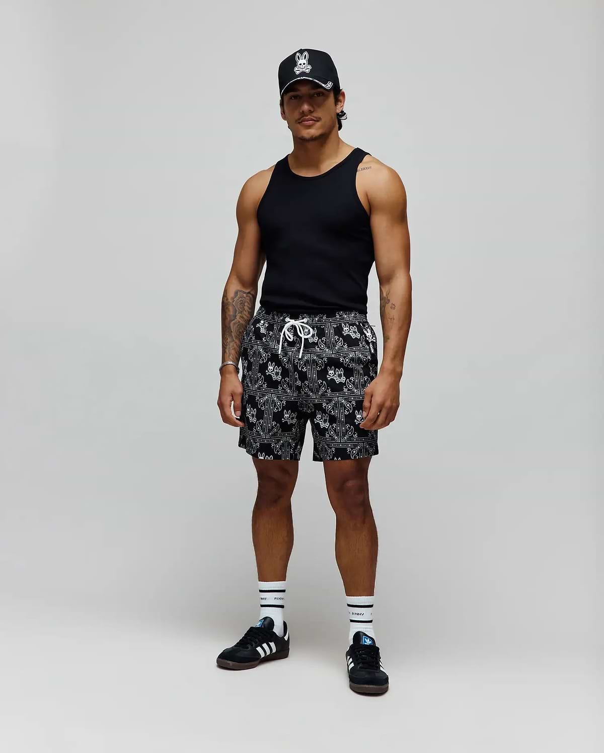 Black tank top, black cap with white embroidered Psycho Bunny skull logo, and Mens Luke Swim Trunk in black with white allover bandana Psycho Bunny print, white drawstring, and white crew socks.