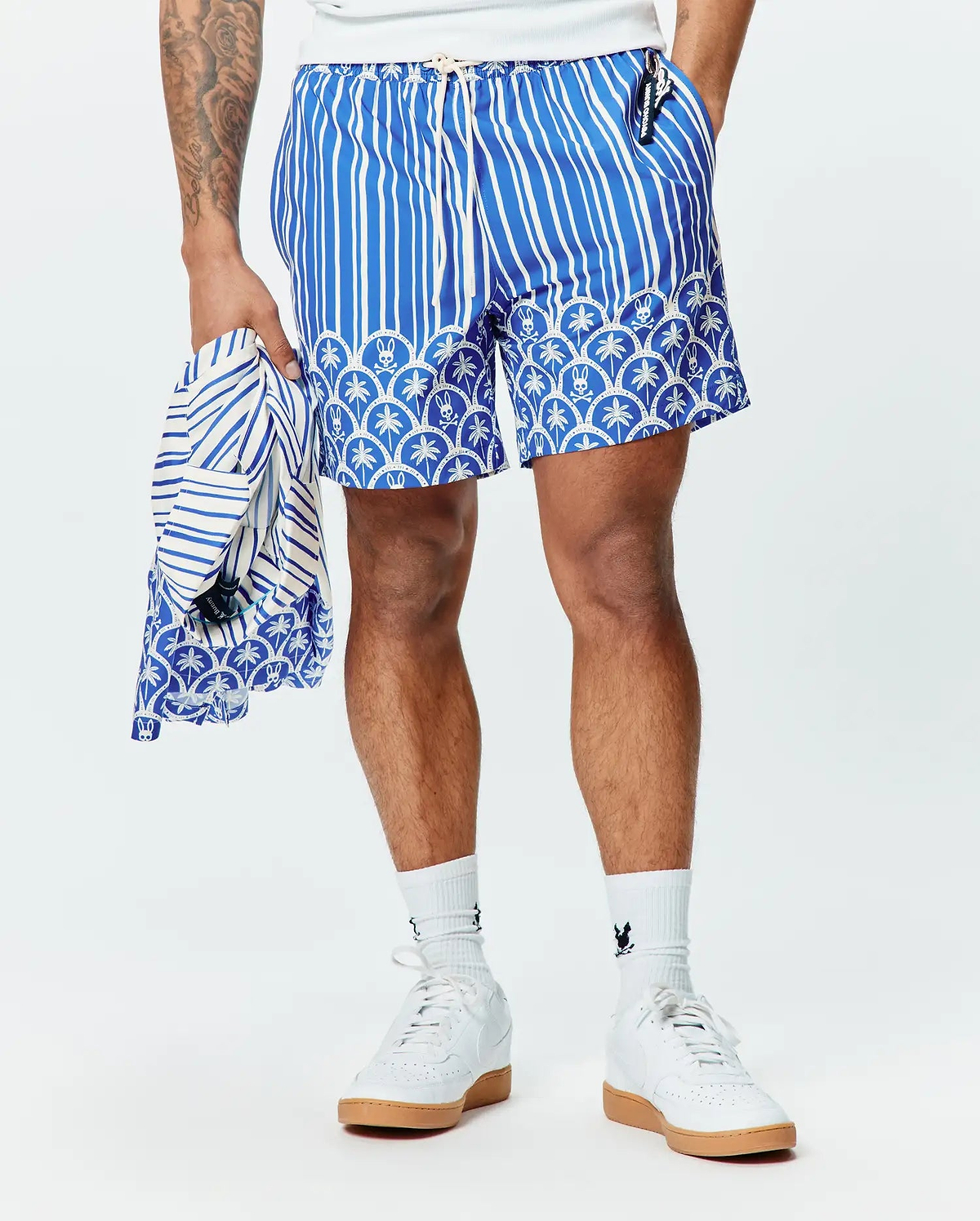 Mens Brad Swim Trunk in blue with white vertical stripes, transitioning to a scalloped pattern featuring palm trees and small Psycho Bunny skull logos. Relaxed fit, smooth fabric.