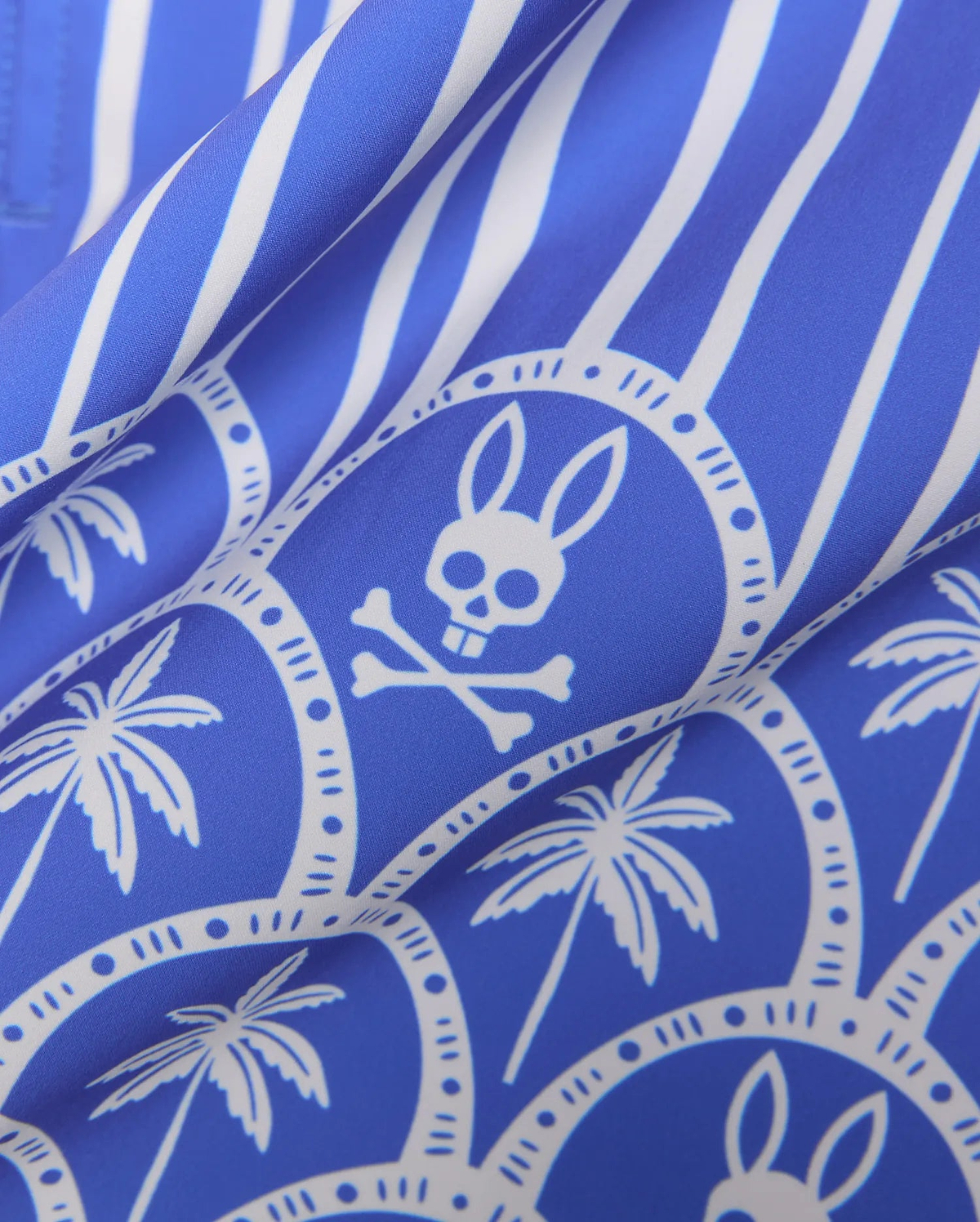 Mens Brad Swim Trunk in royal blue with white vertical stripes, an all-over print featuring white palm trees, and the iconic Psycho Bunny skull logo within decorative circles on a smooth woven fabric.