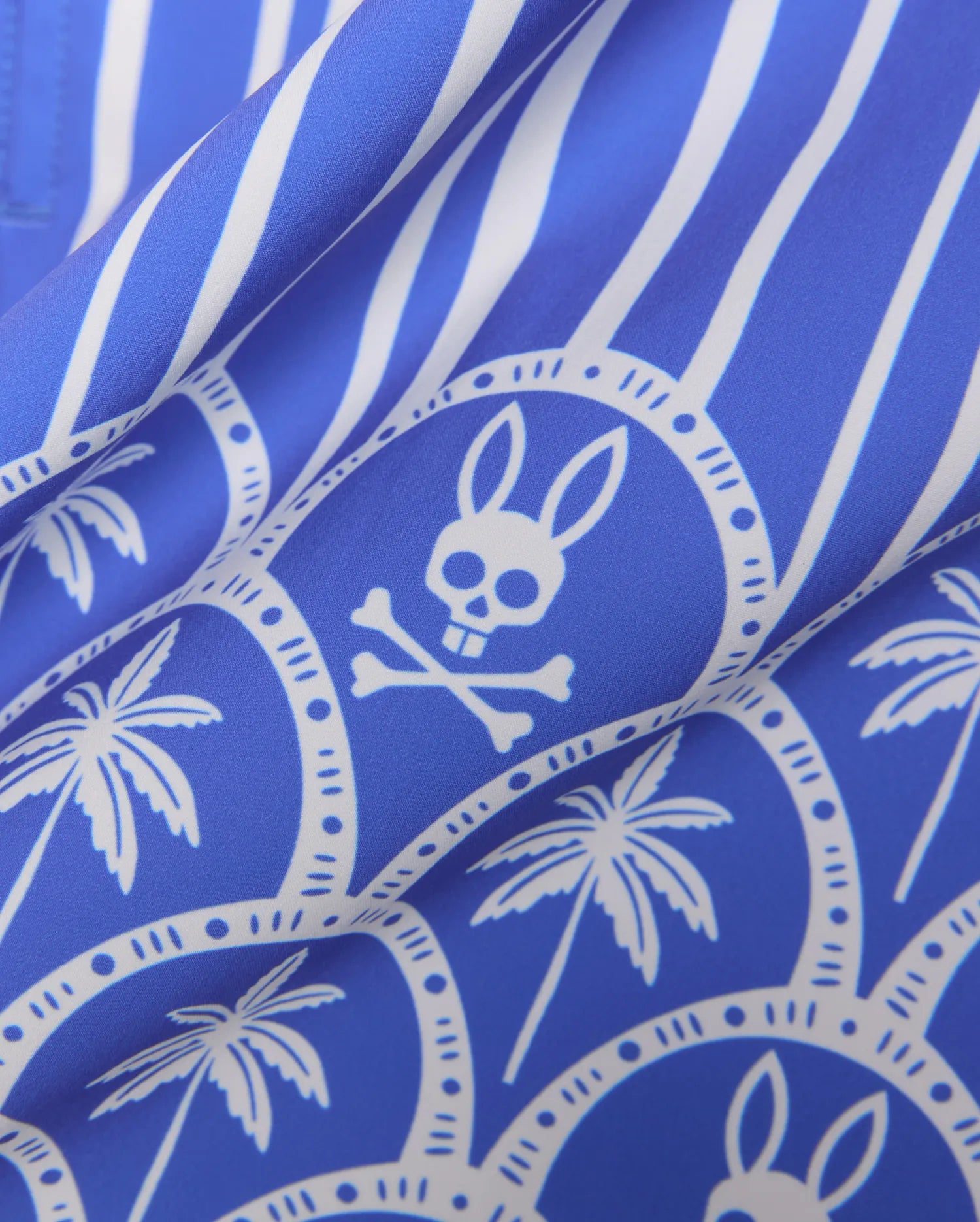 Mens Brad Swim Trunk in royal blue with white vertical stripes, an all-over print featuring white palm trees, and the iconic Psycho Bunny skull logo within decorative circles on a smooth woven fabric.