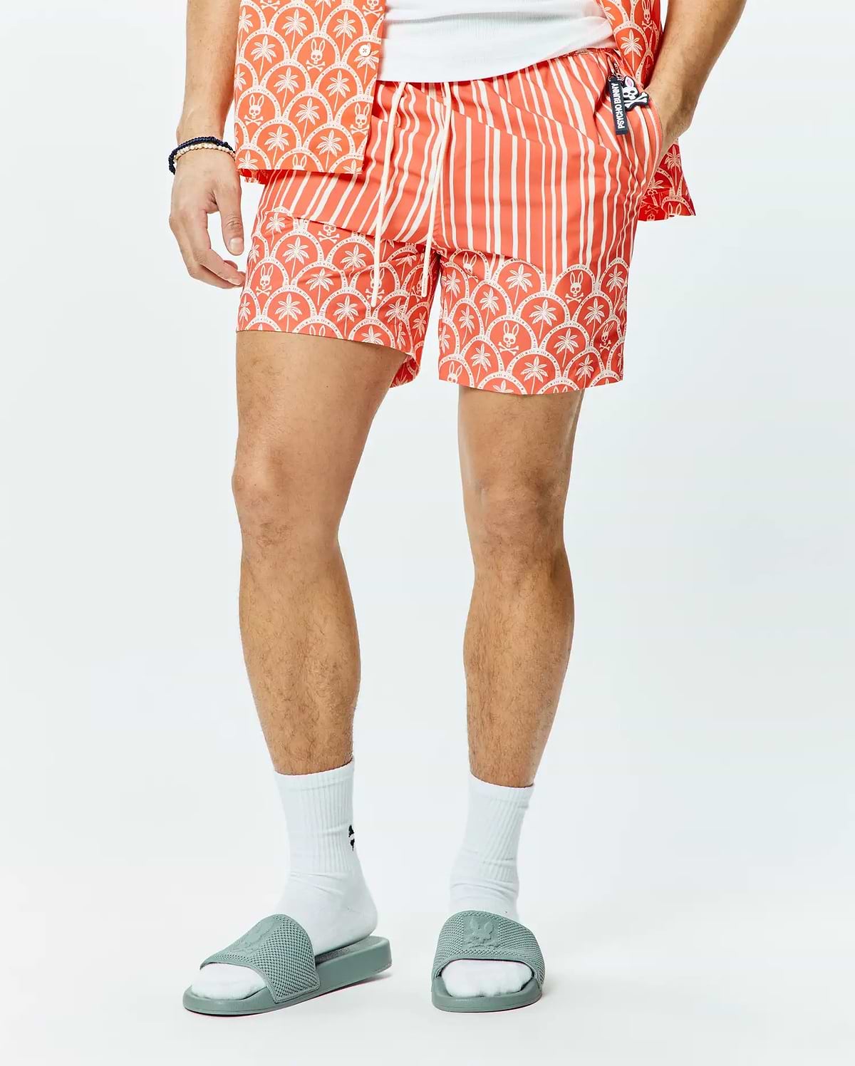 Mens Brad Swim Trunk, a coral orange classic fit swim trunk with white vertical stripes, a white palm tree and Psycho Bunny skull pattern border, and a small Psycho Bunny logo on the drawstring.
