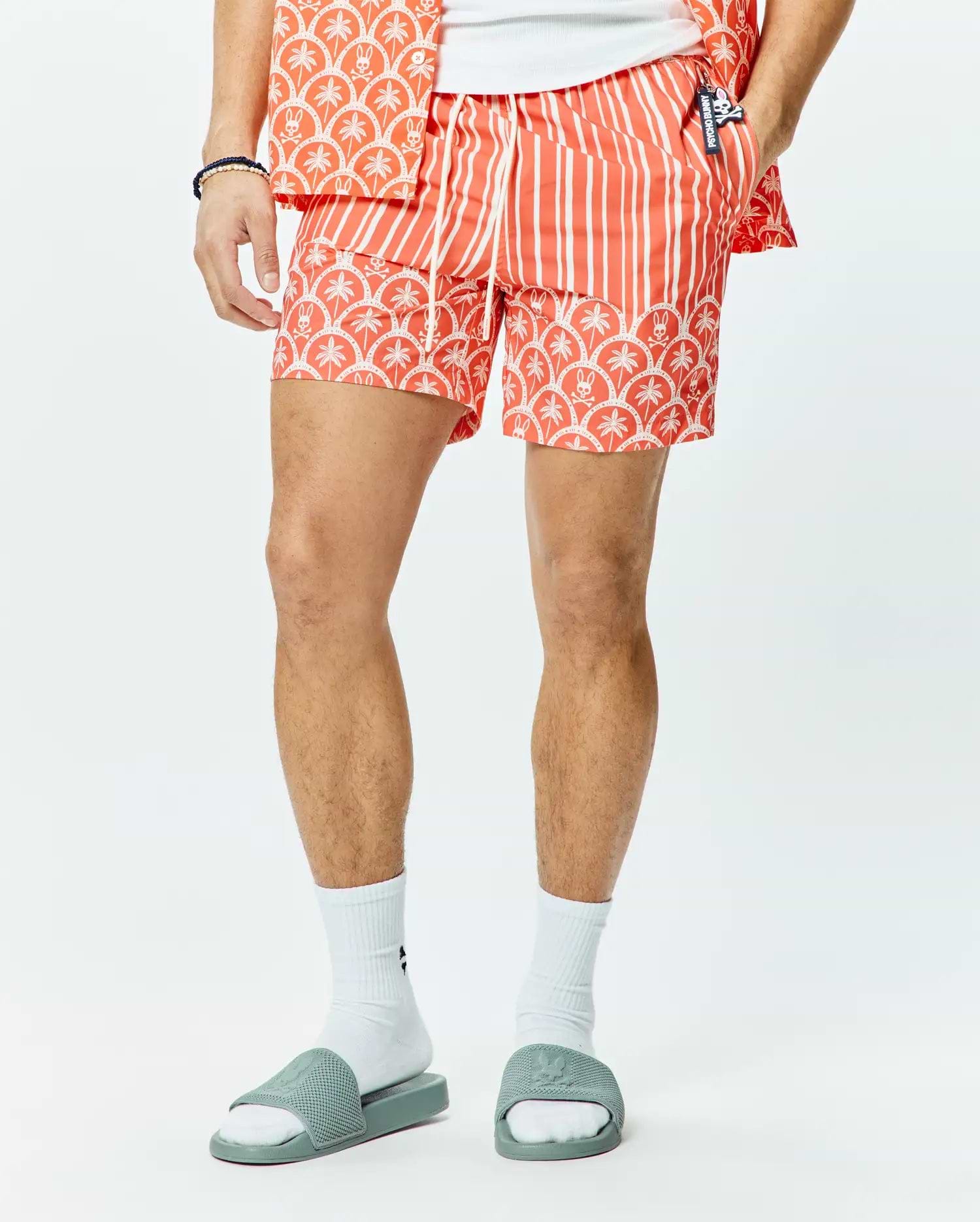 Mens Brad Swim Trunk, a coral orange classic fit swim trunk with white vertical stripes, a white palm tree and Psycho Bunny skull pattern border, and a small Psycho Bunny logo on the drawstring.