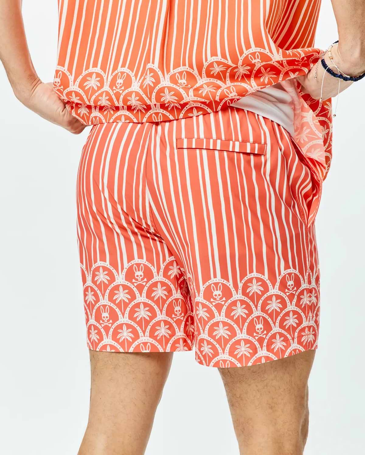Mens Brad Swim Trunk, a relaxed fit coral swim trunk with bold white vertical stripes and a distinctive scalloped hem featuring white palm trees and Psycho Bunny skull logos.