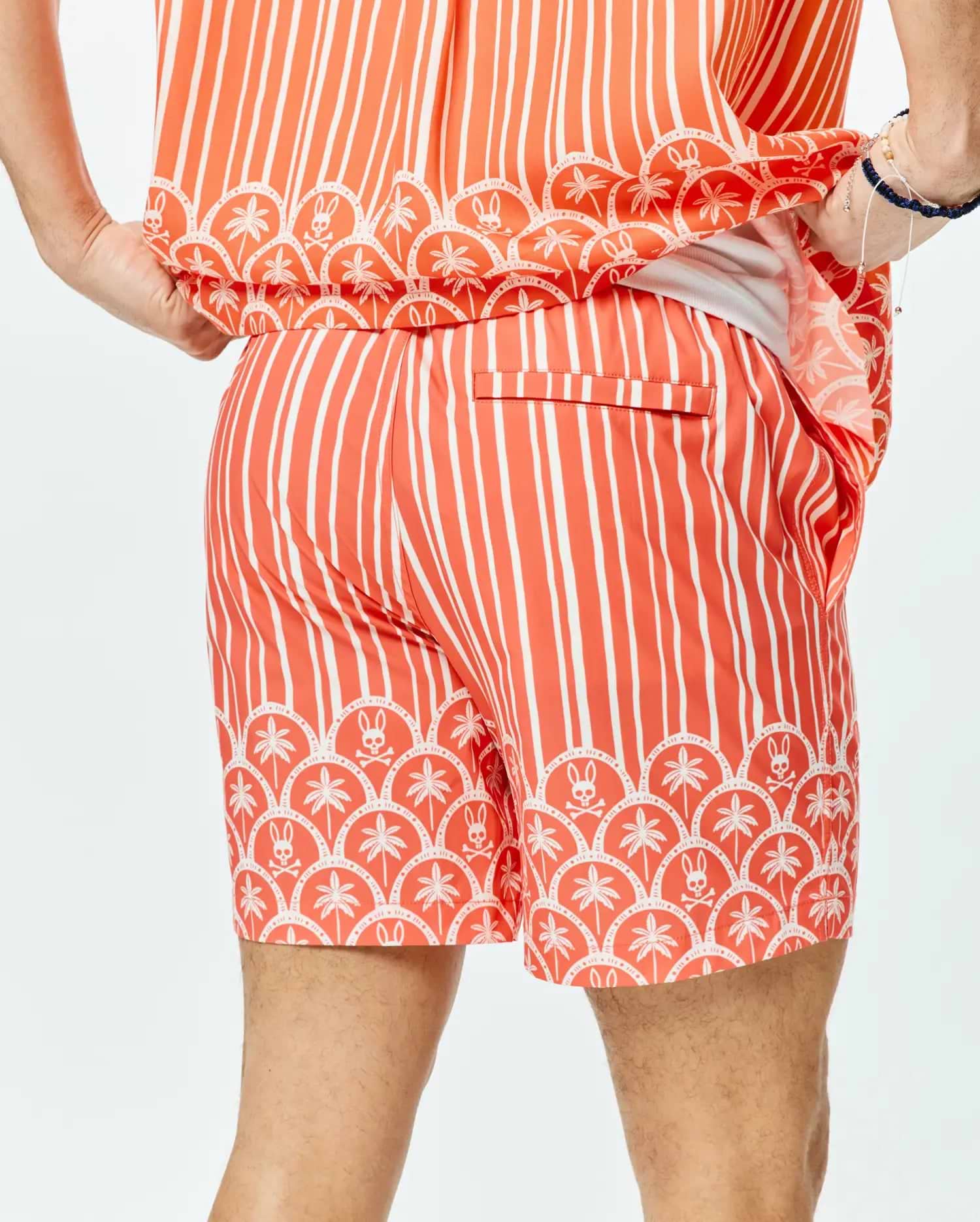 Mens Brad Swim Trunk, a relaxed fit coral swim trunk with bold white vertical stripes and a distinctive scalloped hem featuring white palm trees and Psycho Bunny skull logos.