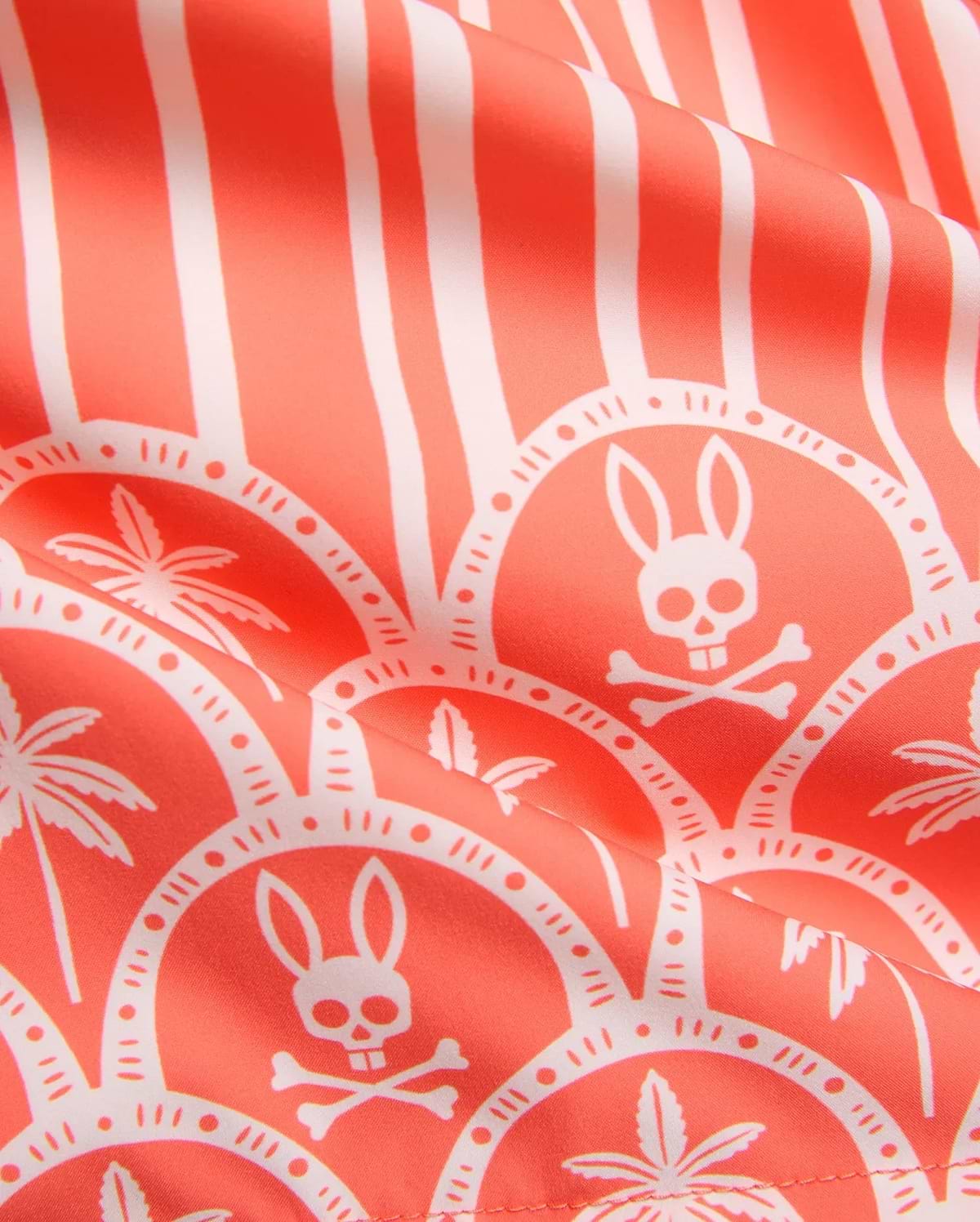 MENS BRAD SWIM TRUNK in vibrant coral with an all-over white print featuring vertical stripes, scalloped patterns, the iconic Psycho Bunny skull logo, and tropical palm trees on smooth fabric.