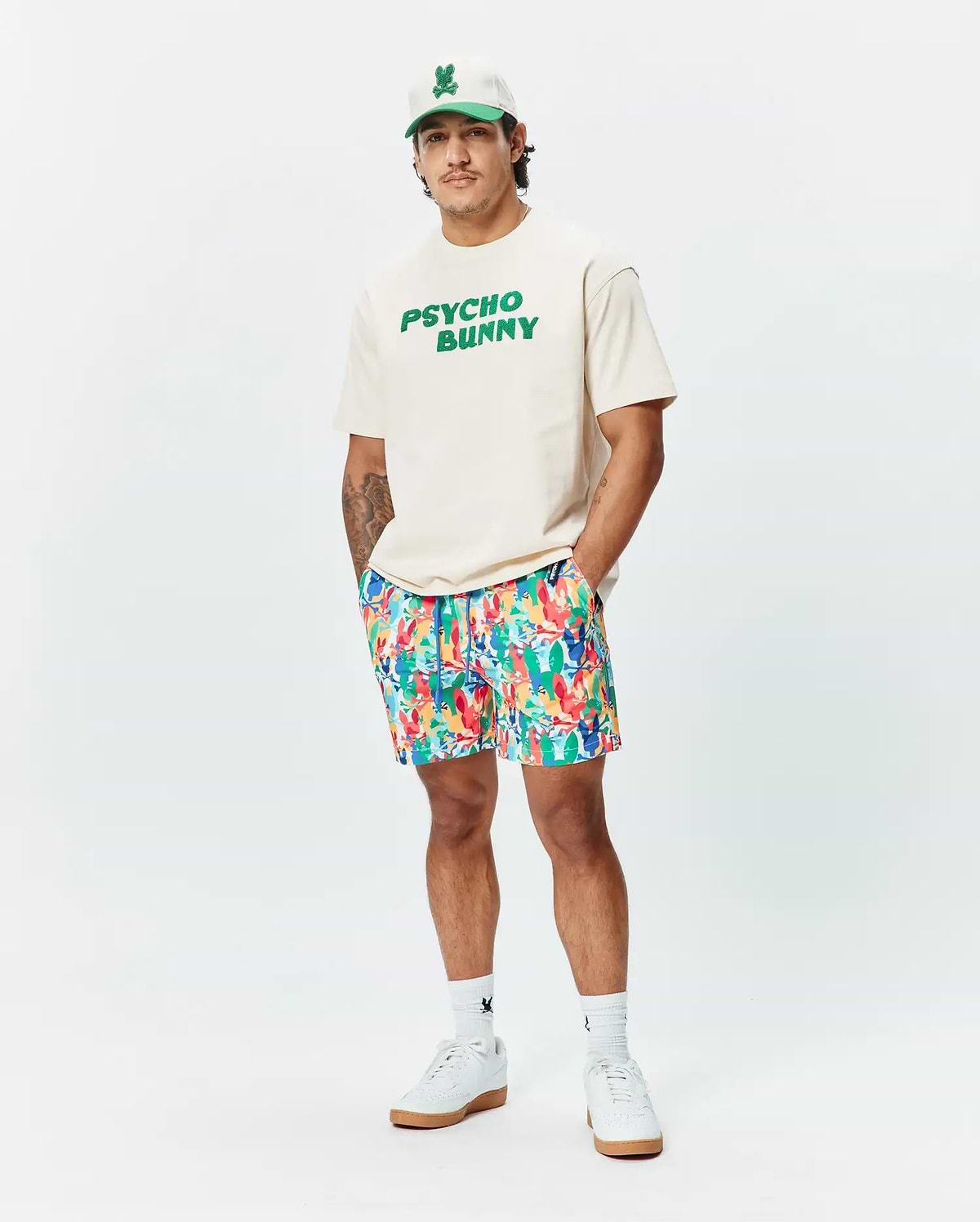 Relaxed fit cream jersey t-shirt with bold green PSYCHO BUNNY chest graphic, vibrant multi-color abstract floral Mens Tanner Swim Trunk, white cap with green bunny skull logo, and white logo socks. (4)