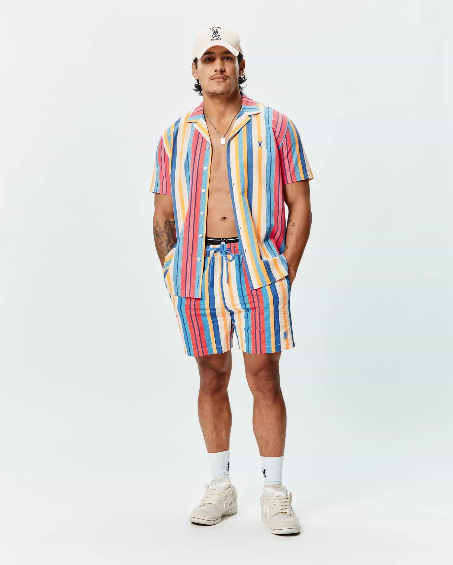 Mens Hubert Swim Trunk in vibrant multi-color vertical stripes (pink, orange, yellow, blue, red), featuring a relaxed fit, textured quick-dry fabric, and a small embroidered Psycho Bunny skull logo on the left leg.