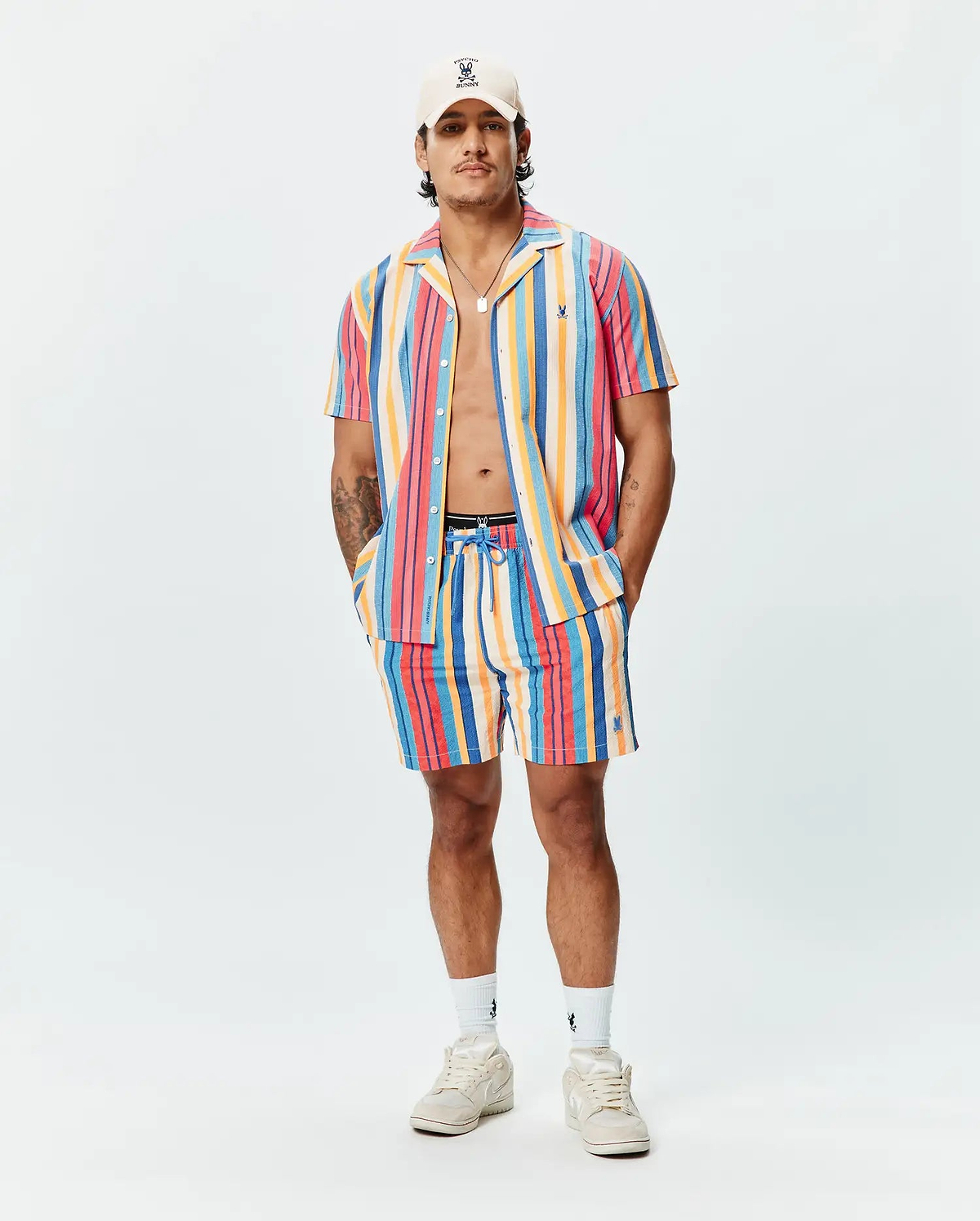 Mens Hubert Swim Trunk in vibrant multi-color vertical stripes (pink, orange, yellow, blue, red), featuring a relaxed fit, textured quick-dry fabric, and a small embroidered Psycho Bunny skull logo on the left leg.