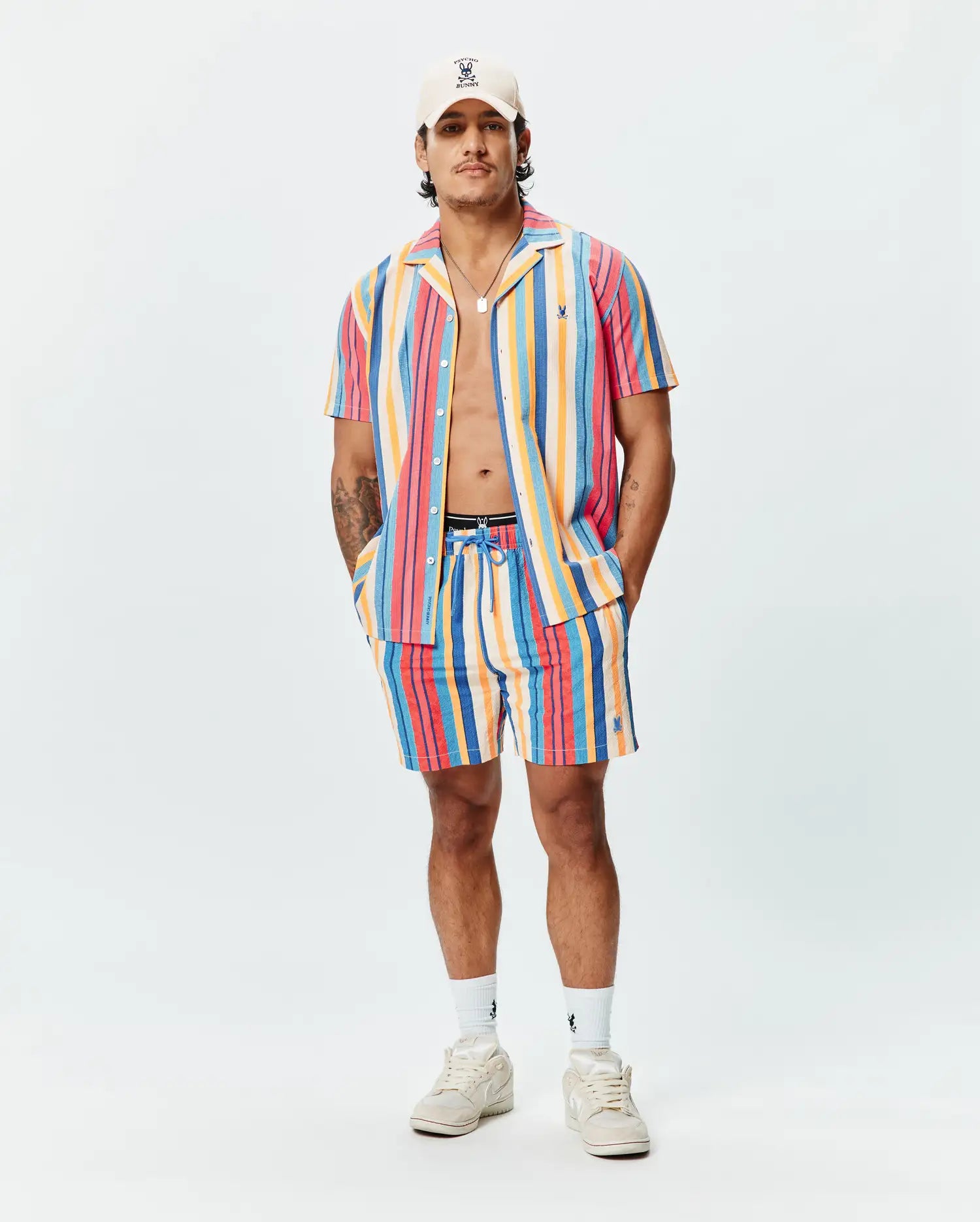 Mens Hubert Swim Trunk in vibrant multi-color vertical stripes (pink, orange, yellow, blue, red), featuring a relaxed fit, textured quick-dry fabric, and a small embroidered Psycho Bunny skull logo on the left leg.
