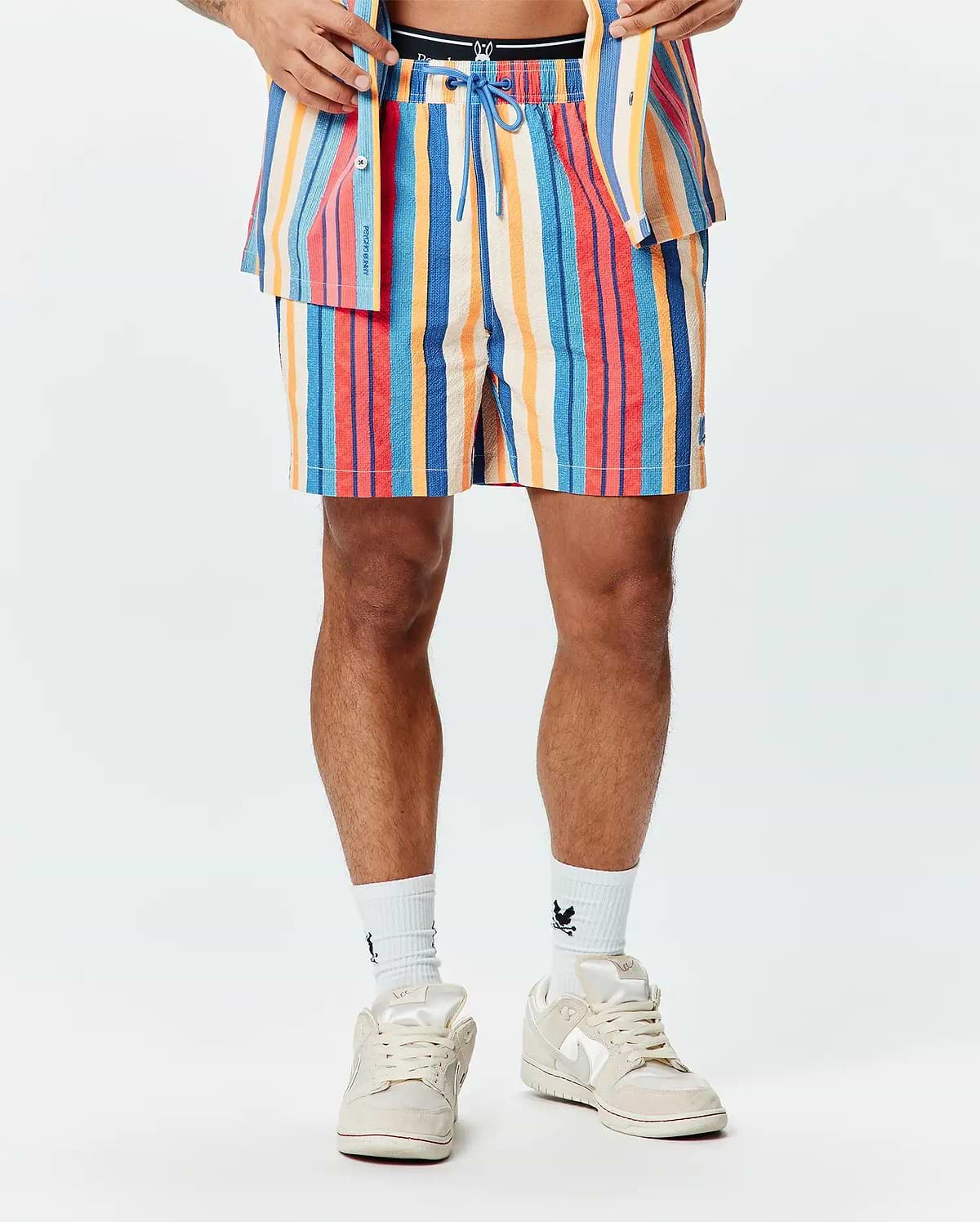 Mens Hubert Swim Trunk, a multi-colored striped swim trunk with blue, red, orange, and cream vertical stripes, featuring a textured fabric, blue drawstring, and a small embroidered Psycho Bunny skull logo on the left leg.