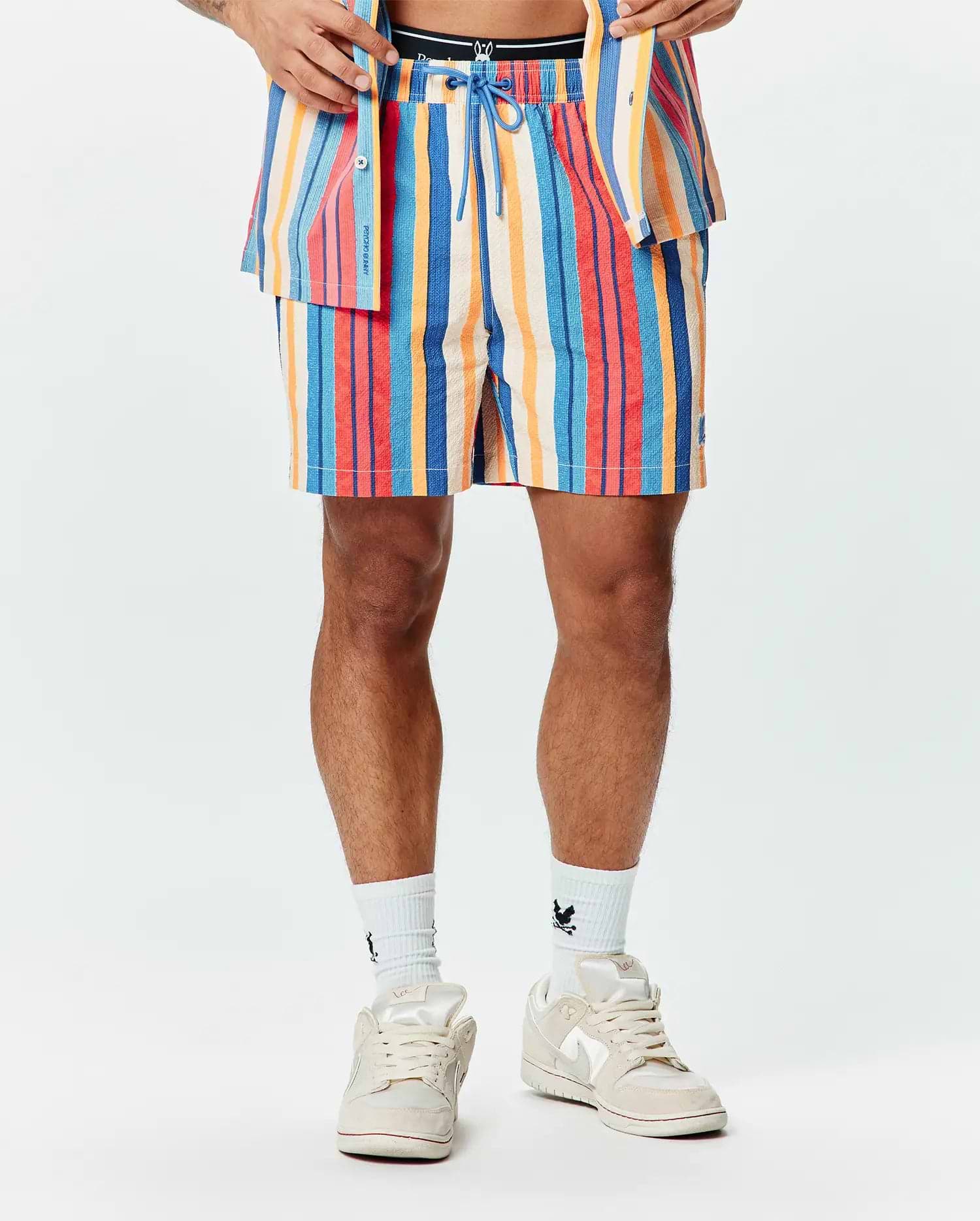 Mens Hubert Swim Trunk, a multi-colored striped swim trunk with blue, red, orange, and cream vertical stripes, featuring a textured fabric, blue drawstring, and a small embroidered Psycho Bunny skull logo on the left leg.