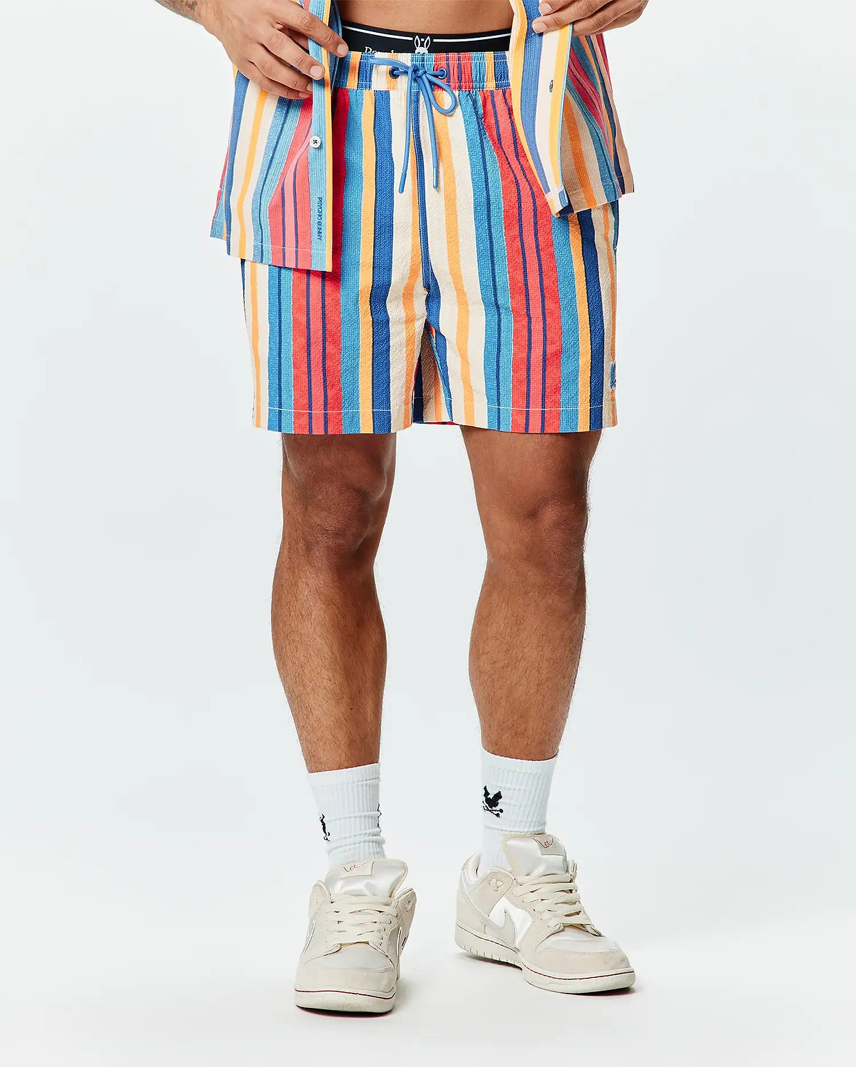 Mens Hubert Swim Trunk, a multi-colored striped swim trunk with blue, red, orange, and cream vertical stripes, featuring a textured fabric, blue drawstring, and a small embroidered Psycho Bunny skull logo on the left leg.