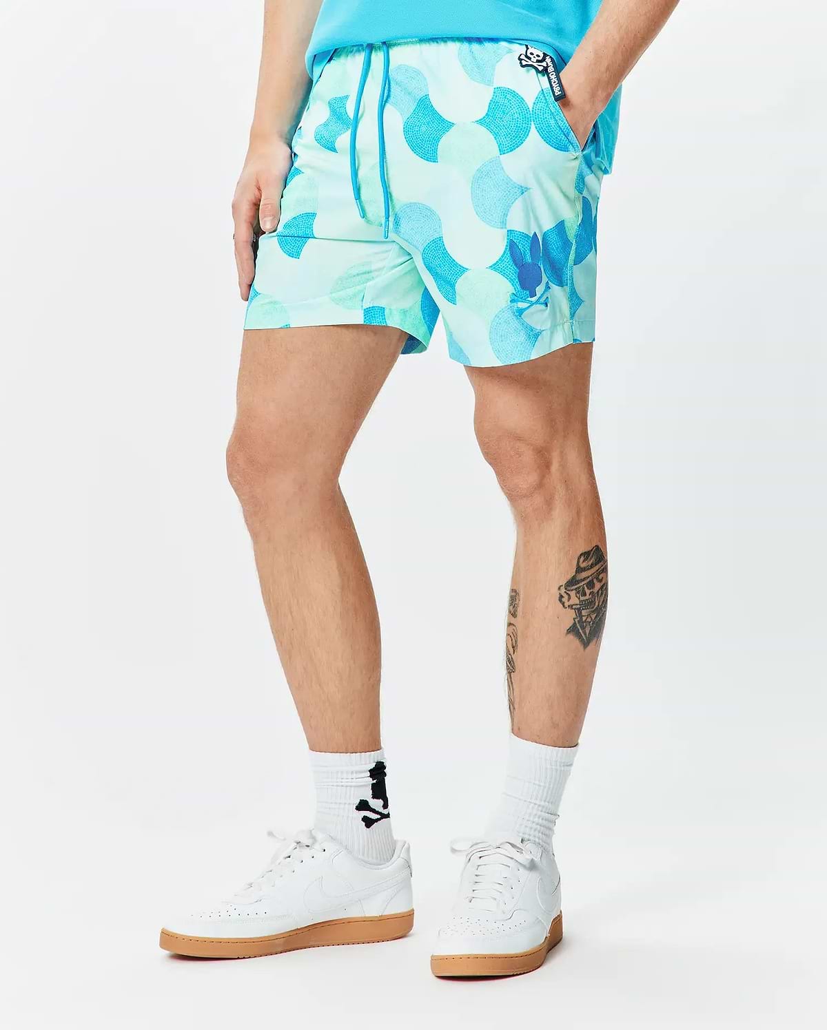 MENS ETHAN SWIM TRUNK 6, vibrant aqua and teal mosaic pattern, slim fit, smooth quick-dry fabric, blue drawstring, small Psycho Bunny logo tag, and a larger blue bunny skull graphic on the left leg. (1)