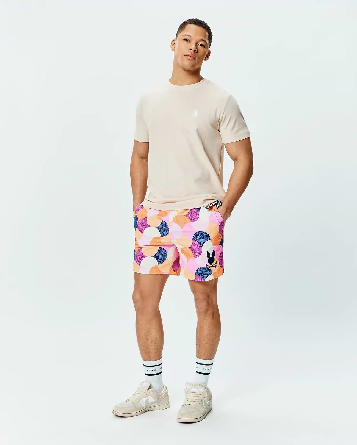 Mens Ethan Swim Trunk 6 with a multi-color abstract geometric pattern, featuring a black Psycho Bunny skull logo. Paired with a classic fit beige jersey tee with a white Psycho Bunny skull logo. (4)