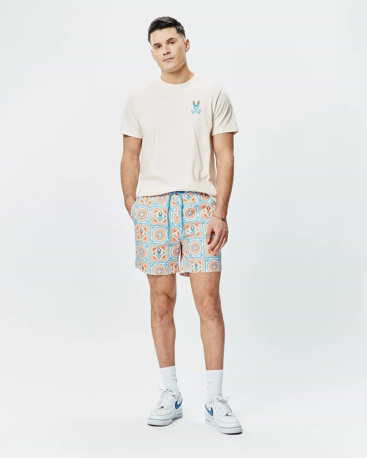 Cream jersey t-shirt with a blue and orange Psycho Bunny logo on the chest, paired with Mens Frank Swim Trunk 6 featuring a blue and orange geometric tile print with integrated bunny skull motifs.
