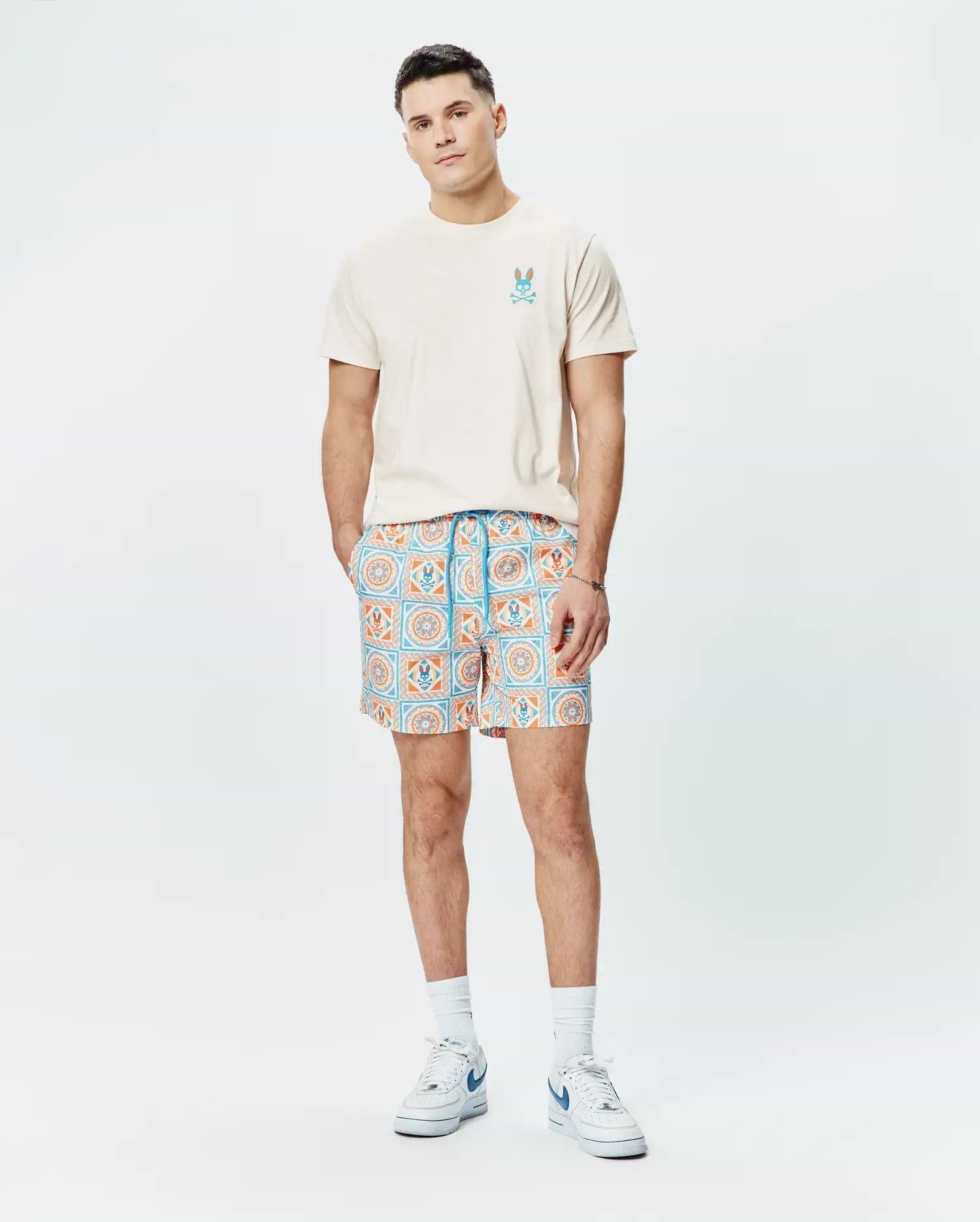 Cream jersey t-shirt with a blue and orange Psycho Bunny logo on the chest, paired with Mens Frank Swim Trunk 6 featuring a blue and orange geometric tile print with integrated bunny skull motifs.