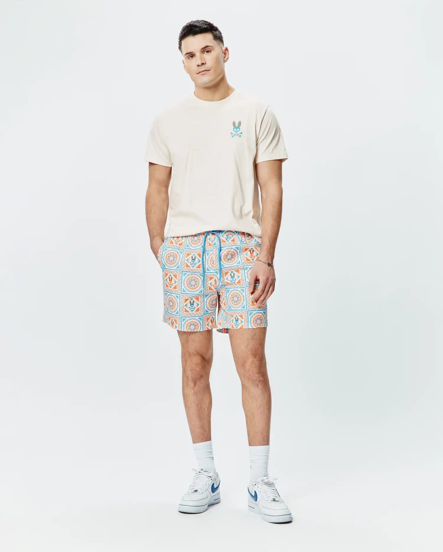 Cream jersey t-shirt with a blue and orange Psycho Bunny logo on the chest, paired with Mens Frank Swim Trunk 6 featuring a blue and orange geometric tile print with integrated bunny skull motifs.