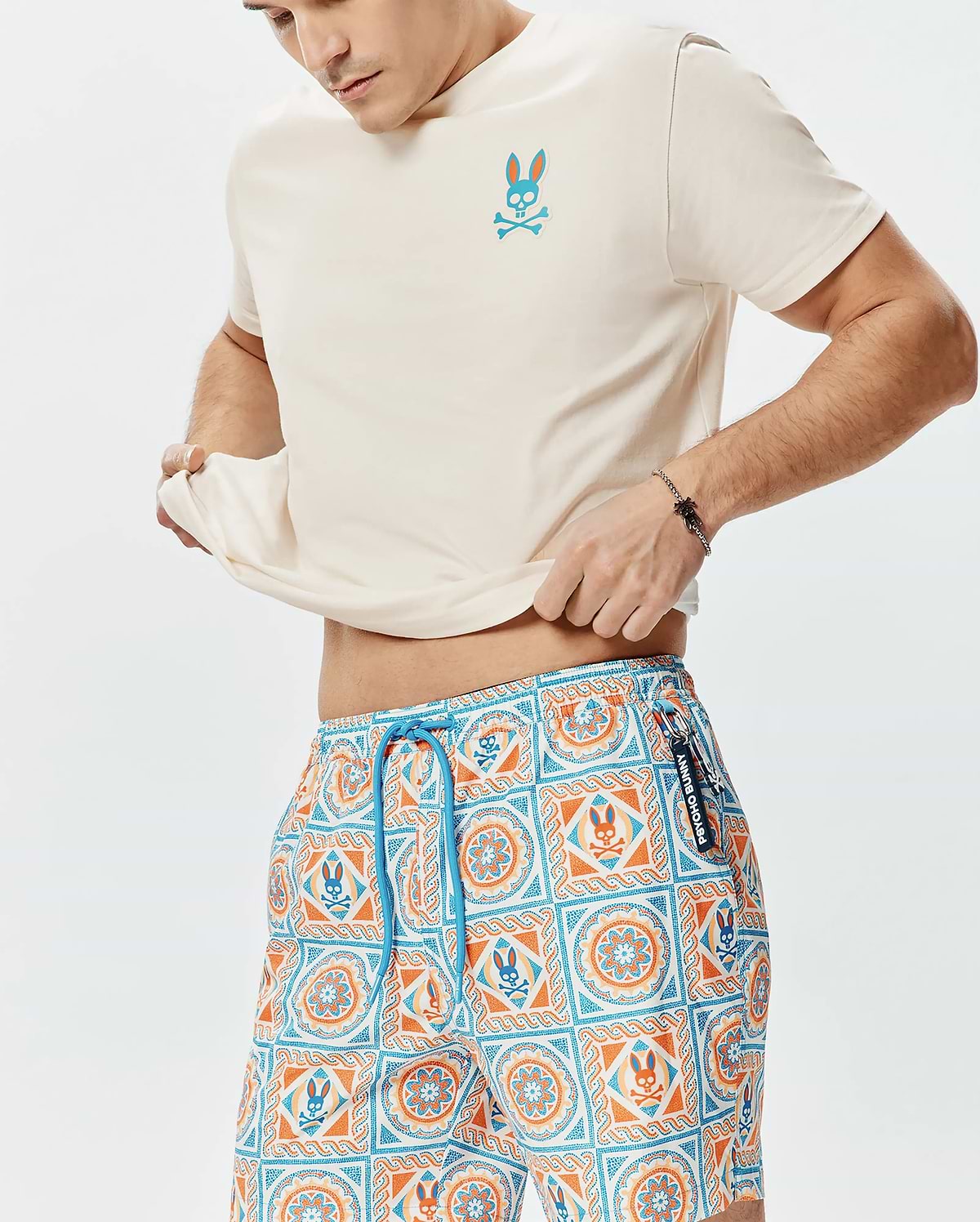 Cream jersey t-shirt with a small embroidered blue and orange Psycho Bunny skull logo on the chest, paired with Mens Frank Swim Trunk 6 featuring a vibrant blue and orange mosaic pattern with integrated Psycho Bunny logos and a blue drawstring.