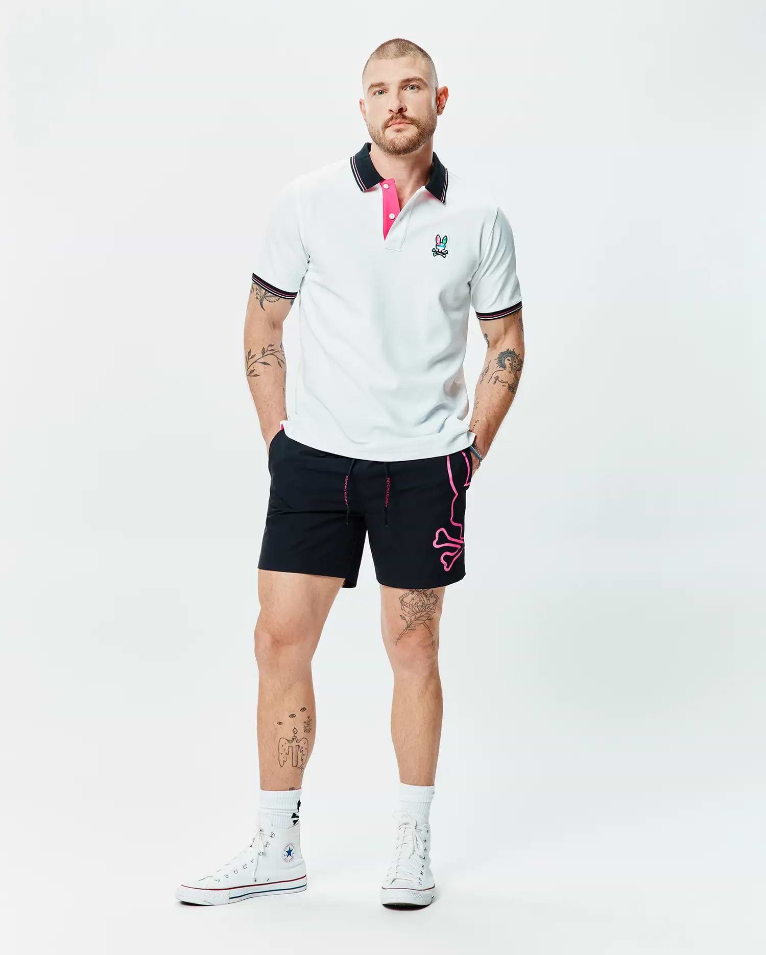 White pique polo with black and pink striped collar, embroidered Psycho Bunny logo, and black Mens Duncan Bunny Print Swim Trunk featuring a large pink bunny skull graphic and pink drawstring.