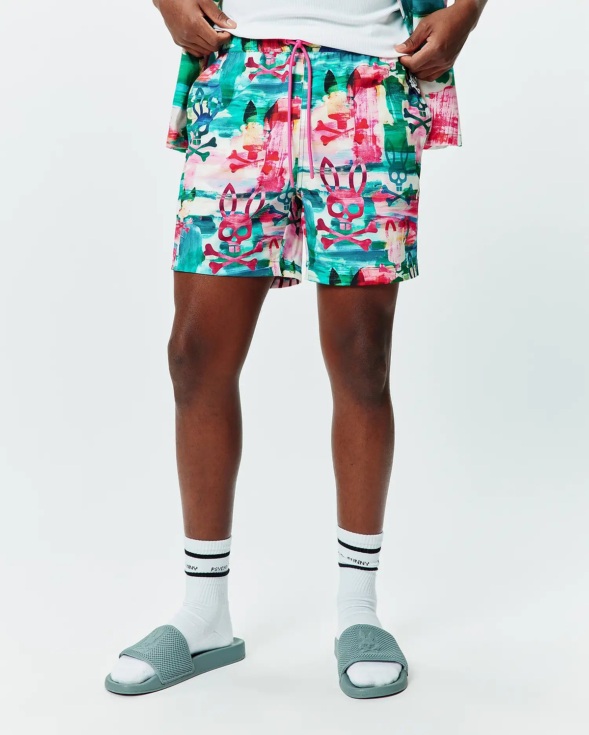 Mens Nate All Over Print Swim Trunk in a vibrant multi-color abstract brushstroke pattern featuring repeated Psycho Bunny skull and crossbones logos, with a pink drawstring, offering a relaxed fit.