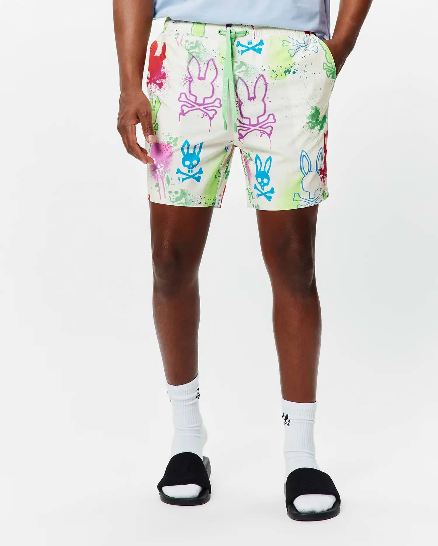 Mens Quintin All Over Print Swim Trunk, a classic fit cream swim trunk with vibrant multi-color Psycho Bunny skull logo and paint splatter all-over print, featuring a neon green drawstring with Psycho Bunny text.