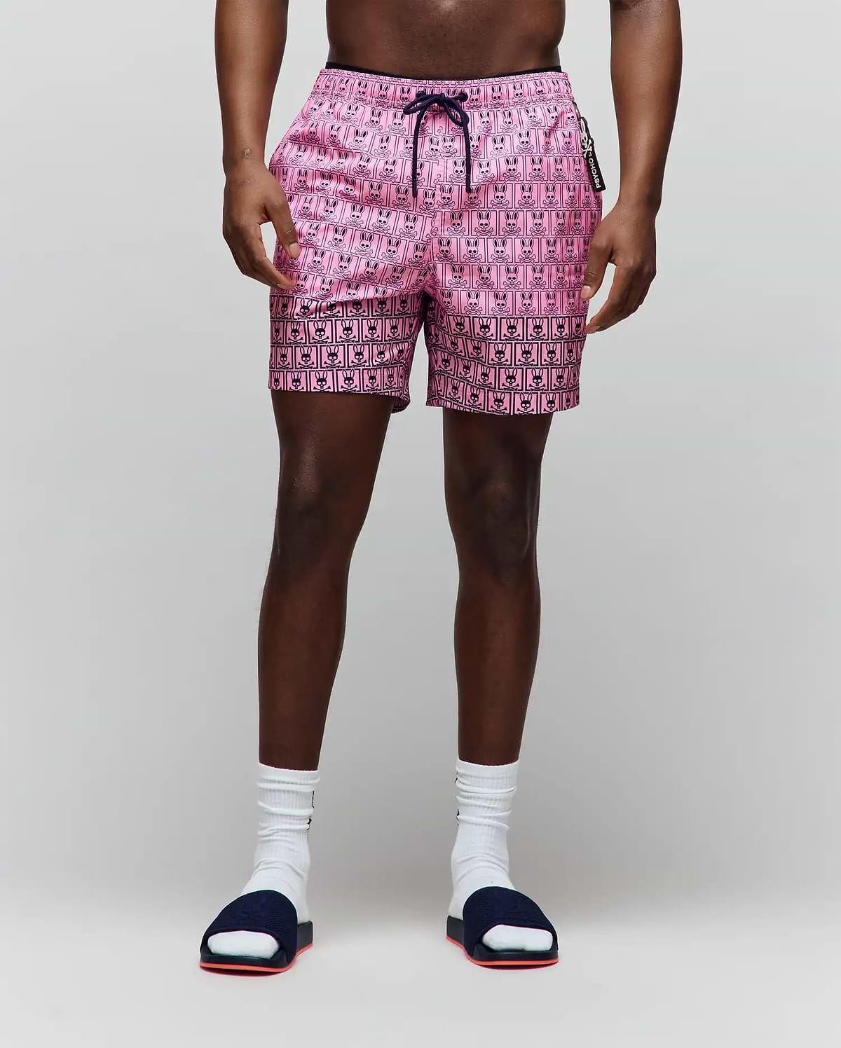 Mens Keon Swim Trunk in pink with an all-over black bunny skull logo pattern, featuring a navy drawstring and a small vertical Psycho Bunny text on the side zipper pull, classic fit. (1)