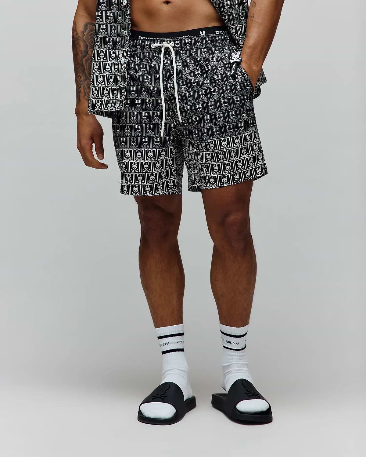 Mens Keon All Over Bunny Swim Trunk, black with a white all-over repeating bunny skull logo pattern, featuring a white drawstring and a black elastic waistband with white Psycho Bunny text, in a relaxed fit.