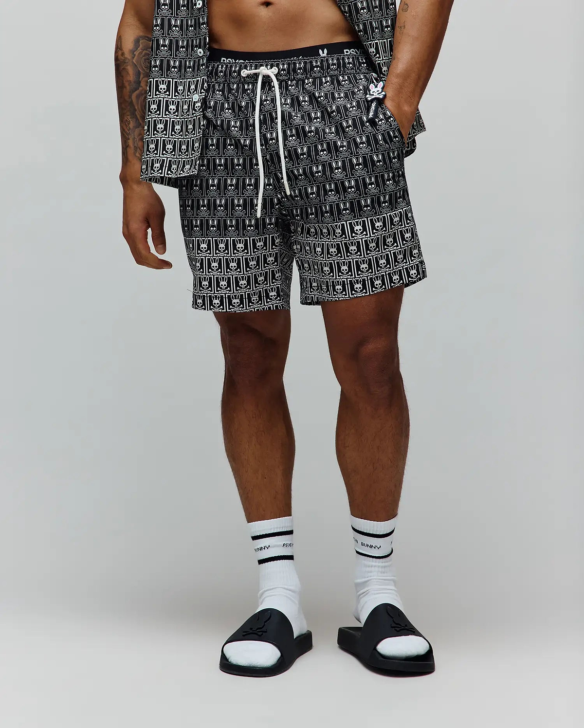 Mens Keon All Over Bunny Swim Trunk, black with a white all-over repeating bunny skull logo pattern, featuring a white drawstring and a black elastic waistband with white Psycho Bunny text, in a relaxed fit.