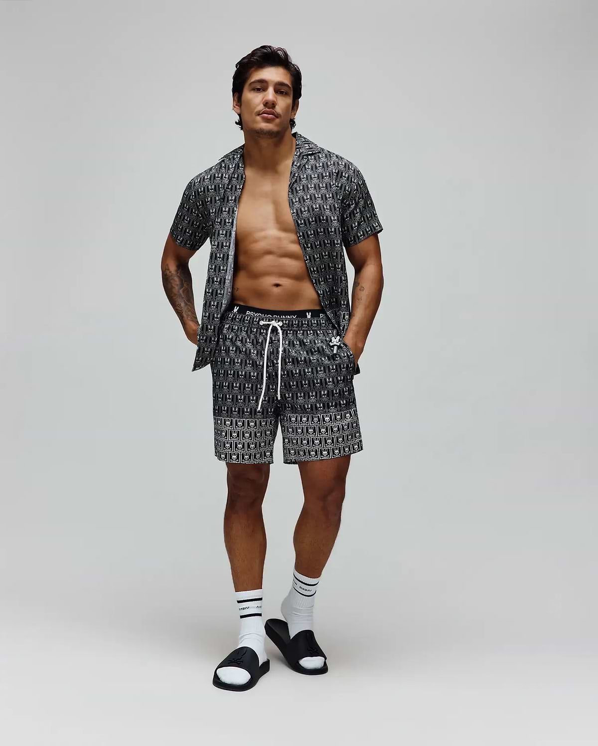 Mens Keon All Over Bunny Swim Trunk and matching short-sleeve shirt in black with a white all-over Psycho Bunny skull logo print, featuring a relaxed fit, smooth fabric, and white drawstring.