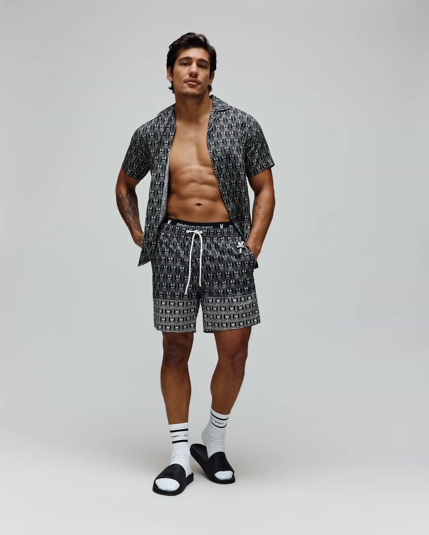 Mens Keon All Over Bunny Swim Trunk and matching short-sleeve shirt in black with a white all-over Psycho Bunny skull logo print, featuring a relaxed fit, smooth fabric, and white drawstring.