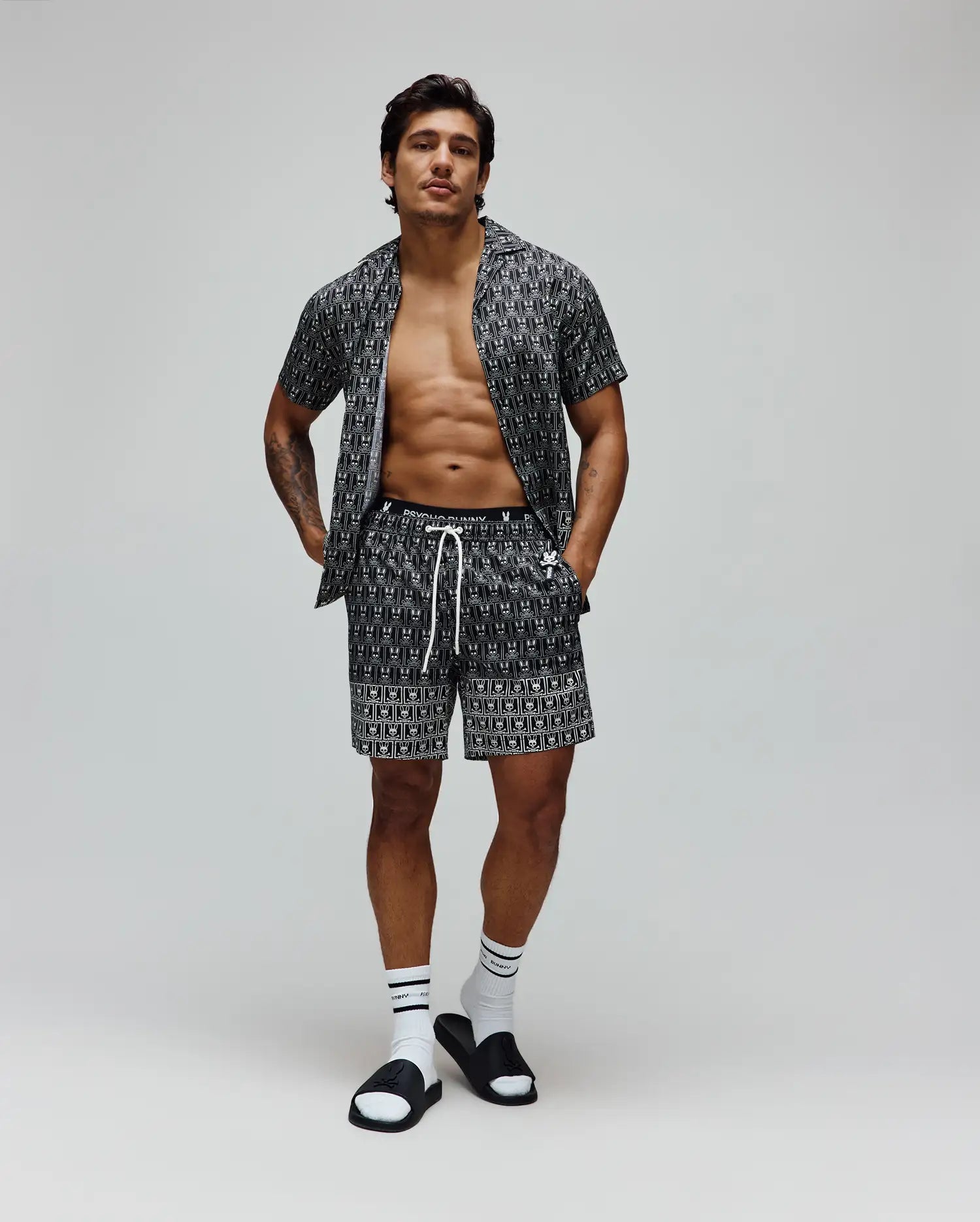 Mens Keon All Over Bunny Swim Trunk and matching short-sleeve shirt in black with a white all-over Psycho Bunny skull logo print, featuring a relaxed fit, smooth fabric, and white drawstring.