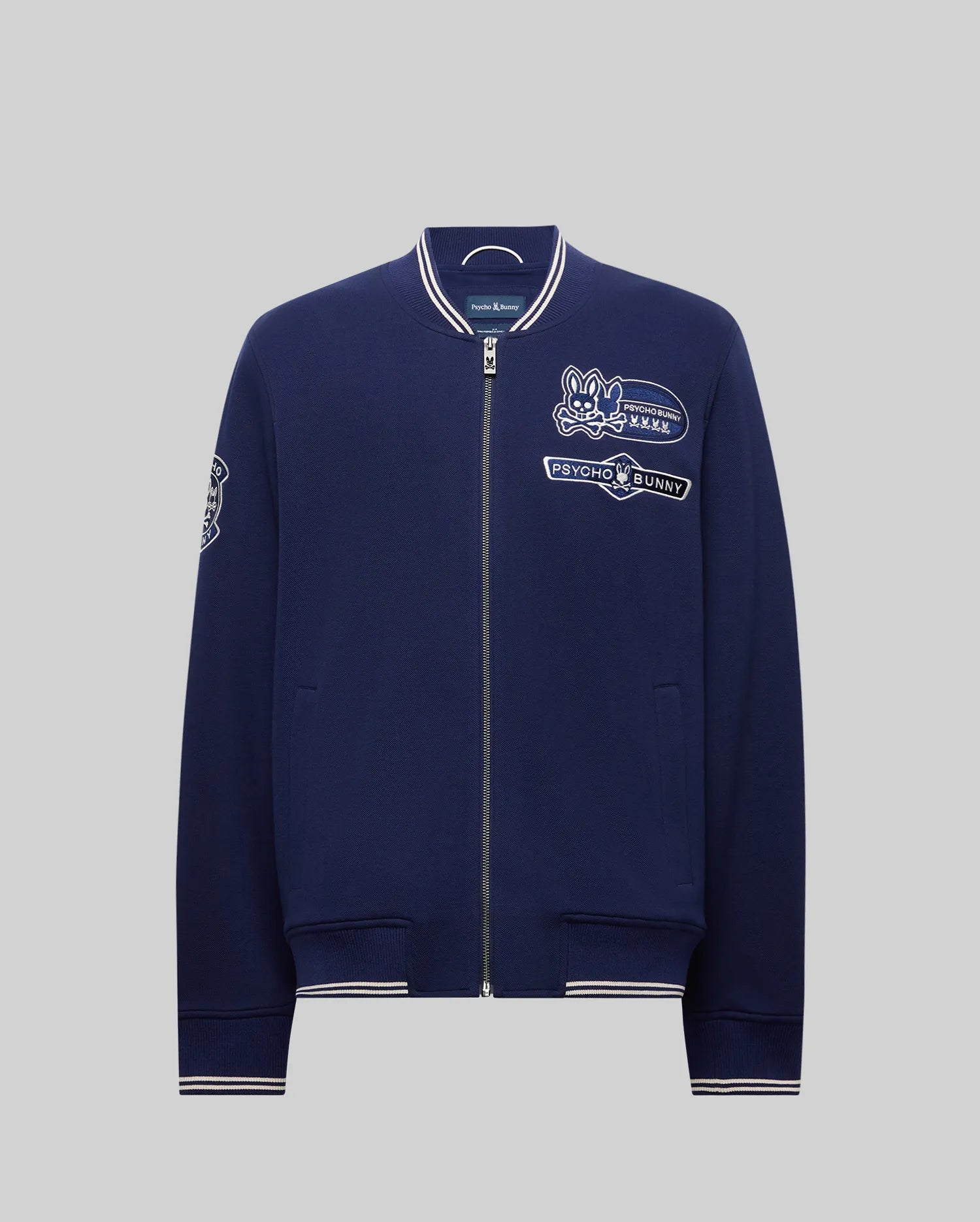 The Psycho Bunny MENS DESMOND PIQUE KNIT BOMBER (MO1100018) is a navy varsity zip-up jacket with white-trimmed collar, cuffs, and hem, embroidered cartoon bunny and "PSYCHO BUNNY" on the chest, plus sleeve designs. (6)