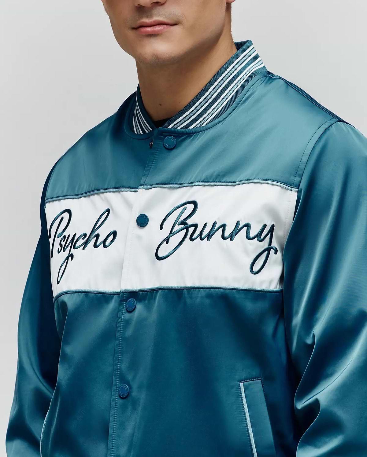 Mens Thomas Satin Bomber in lustrous teal satin, featuring a prominent white chest panel with a large embroidered dark teal Psycho Bunny script logo, and a striped ribbed collar with snap-button closure.