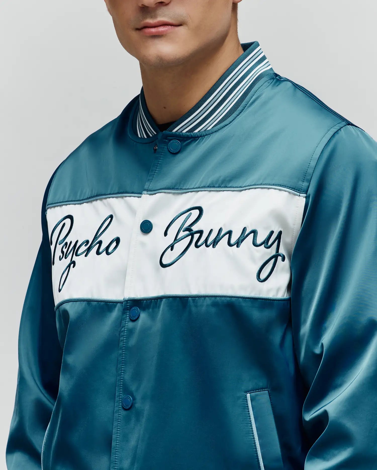 Mens Thomas Satin Bomber in lustrous teal satin, featuring a prominent white chest panel with a large embroidered dark teal Psycho Bunny script logo, and a striped ribbed collar with snap-button closure.