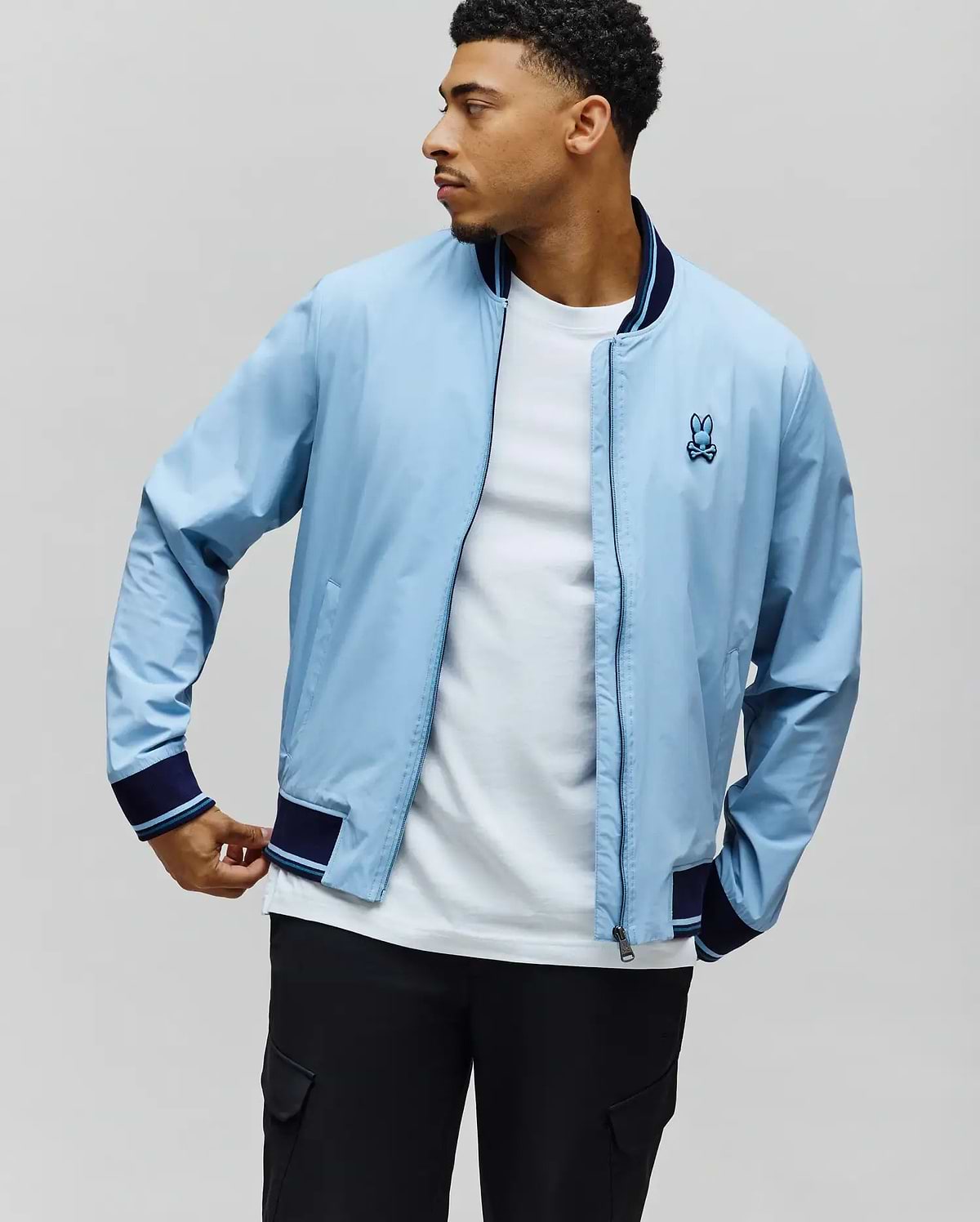 Light blue Mens Aldrin Bomber, classic fit, with navy and light blue striped ribbed collar, cuffs, and hem. Features a small embroidered light blue Psycho Bunny skull logo on the left chest. (1)