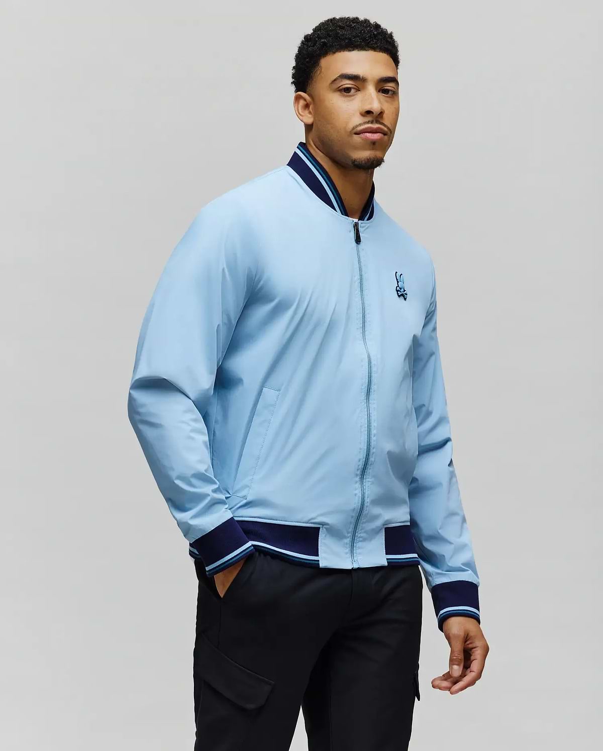 Mens Aldrin Bomber in light blue with navy and light blue striped ribbed collar, cuffs, and hem, featuring a full zip front and an embroidered Psycho Bunny skull logo on the left chest, classic fit. (5)