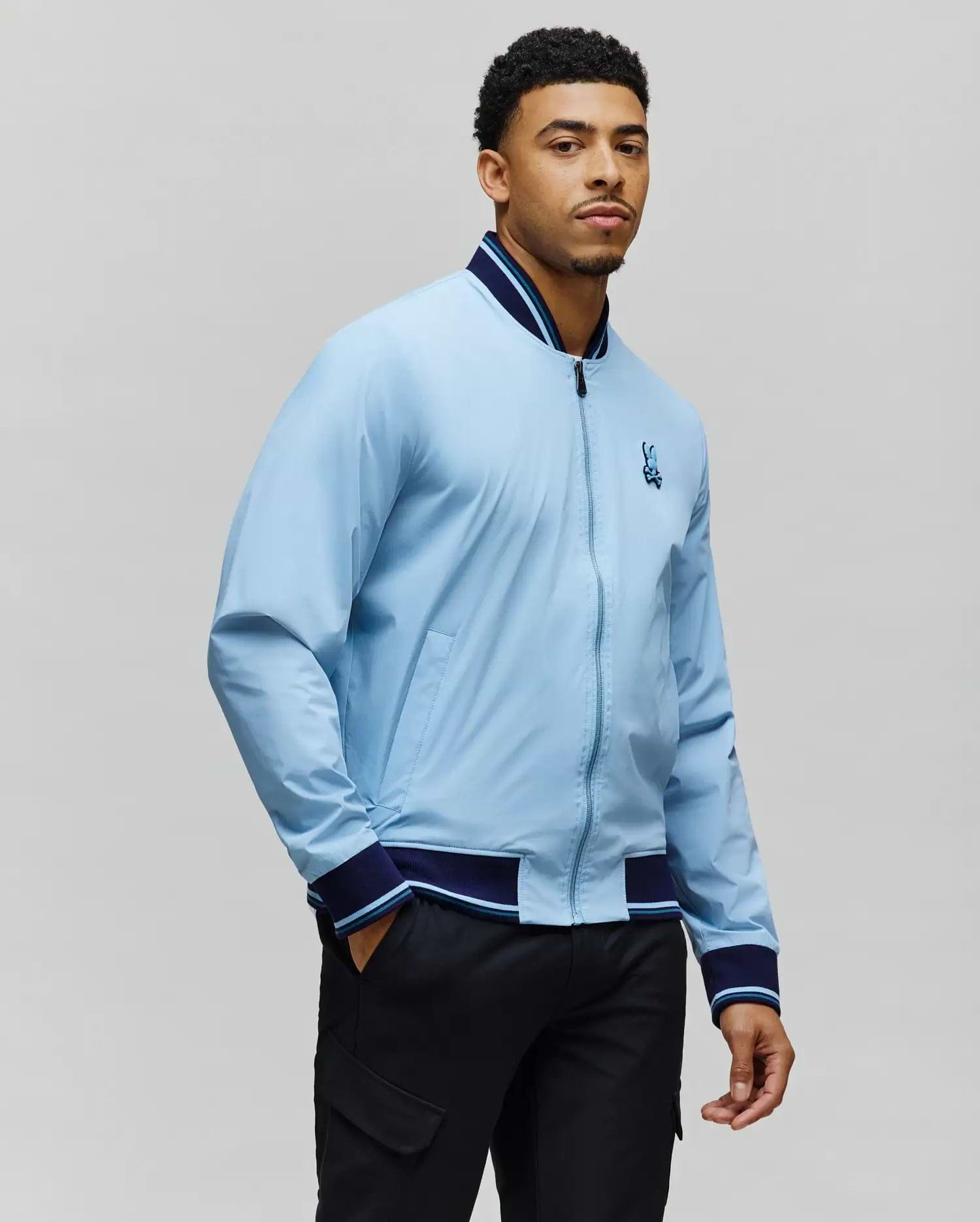 Mens Aldrin Bomber in light blue with navy and light blue striped ribbed collar, cuffs, and hem, featuring a full zip front and an embroidered Psycho Bunny skull logo on the left chest, classic fit. (5)
