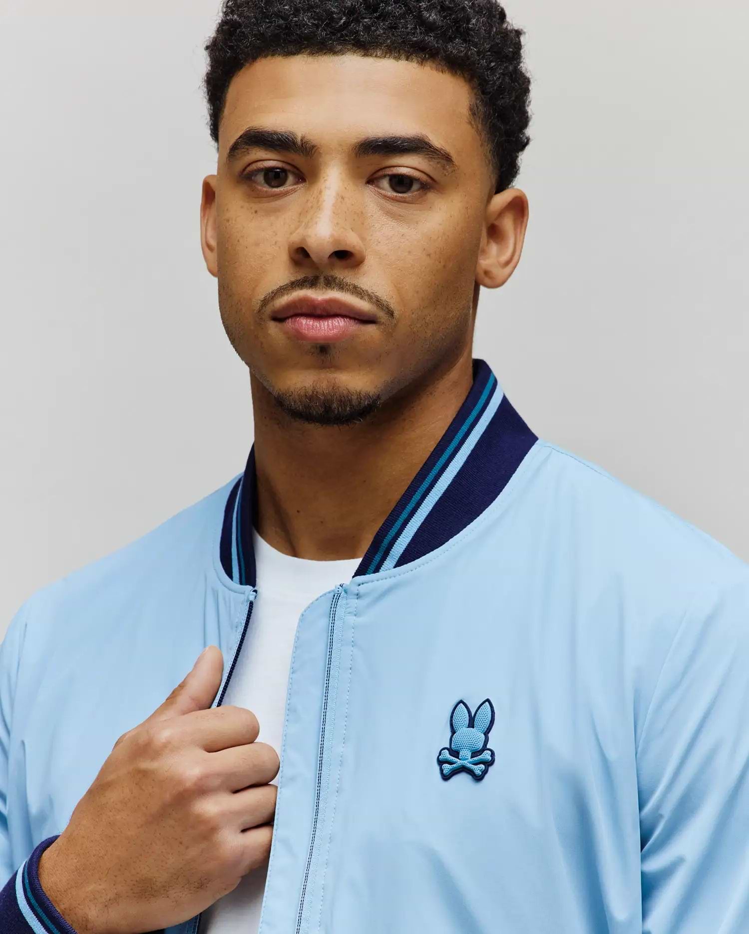 Light blue Mens Aldrin Bomber jacket with a classic fit, featuring a smooth texture, navy and light blue striped ribbed collar and cuffs, and an embroidered Psycho Bunny skull logo on the left chest. (2)