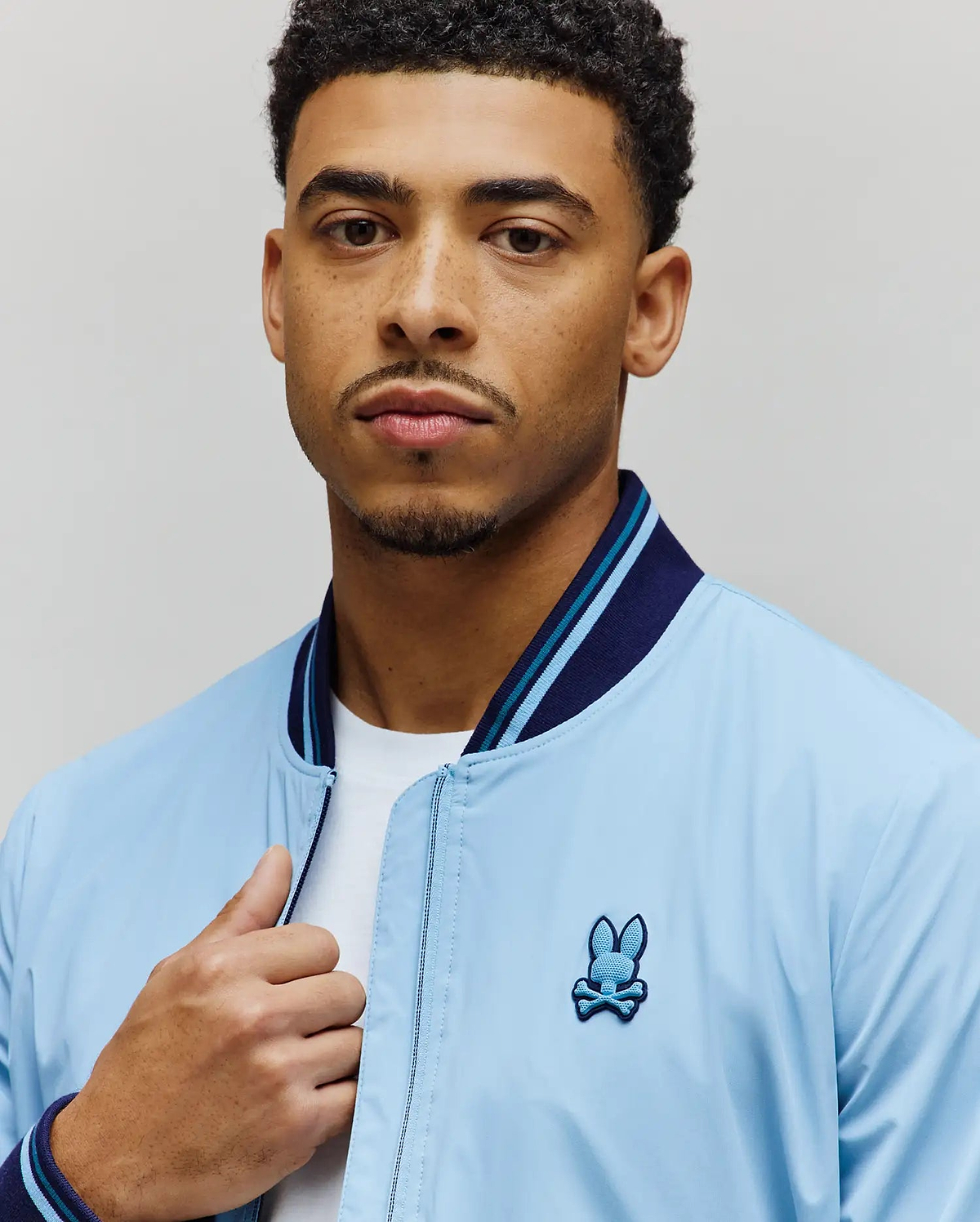 Light blue Mens Aldrin Bomber jacket with a classic fit, featuring a smooth texture, navy and light blue striped ribbed collar and cuffs, and an embroidered Psycho Bunny skull logo on the left chest. (2)