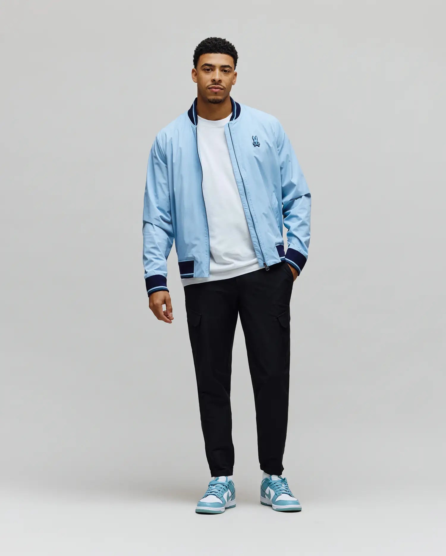 Sky Blue Mens Aldrin Bomber jacket, relaxed fit, with navy/white striped ribbed collar, cuffs, hem, and an embroidered Psycho Bunny skull logo on the left chest. (6)