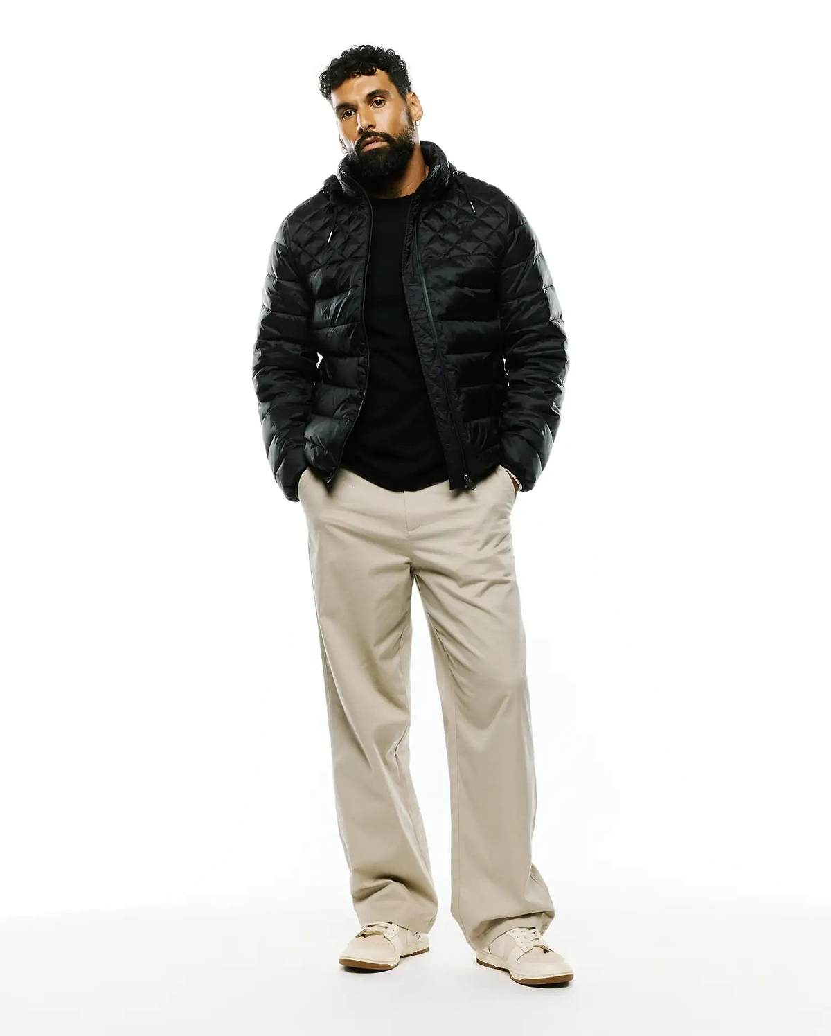 Black Mens Emerik Hooded Puffer Jacket with a classic fit, featuring diamond quilting and a shiny finish. Paired with a black jersey t-shirt and relaxed khaki wide-leg pants.