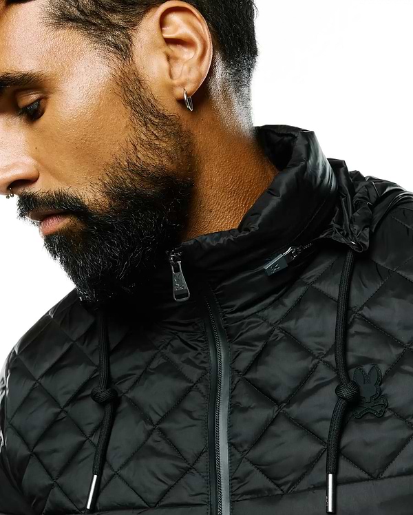 Black Mens Emerik Hooded Puffer Jacket with a quilted diamond pattern, featuring a small black rubberized Psycho Bunny skull logo on the left chest, silver-toned zipper pull and drawstring tips.