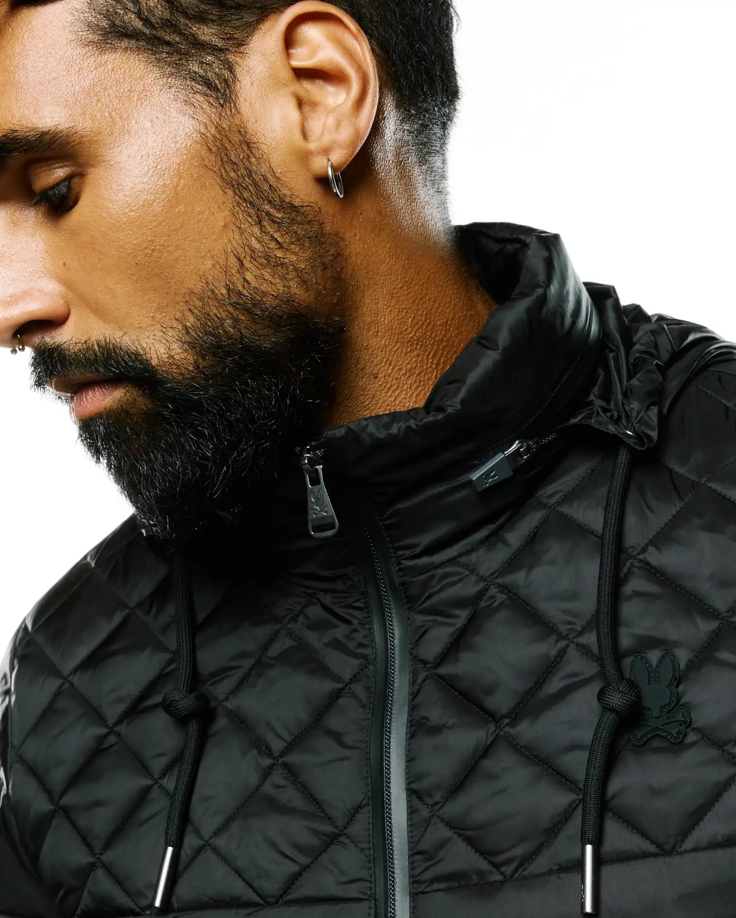 Black Mens Emerik Hooded Puffer Jacket with a quilted diamond pattern, featuring a small black rubberized Psycho Bunny skull logo on the left chest, silver-toned zipper pull and drawstring tips.
